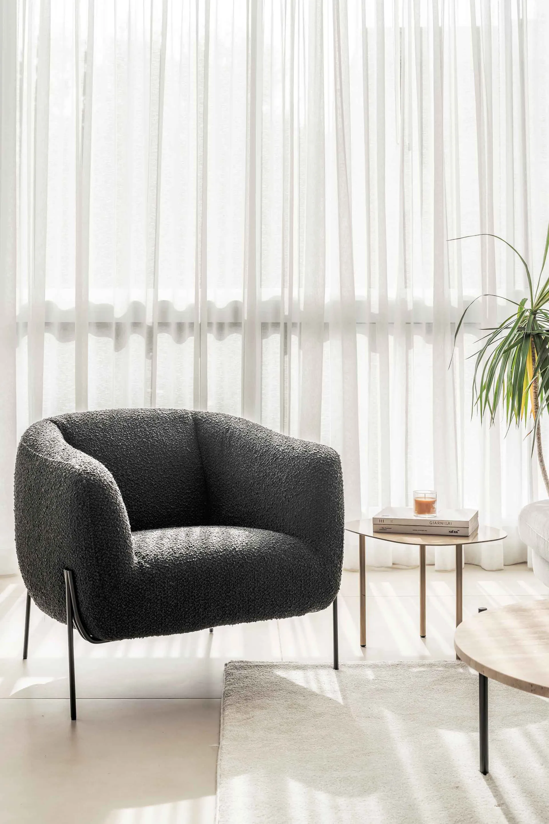 A modern living room with a comfortable fabric chair, white sheer curtains, and a small wooden table with a candle and book.