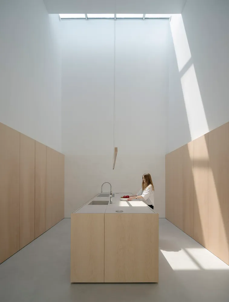 Person preparing food in a minimalist modern kitchen with wood cabinets, white countertops, and overhead lighting.