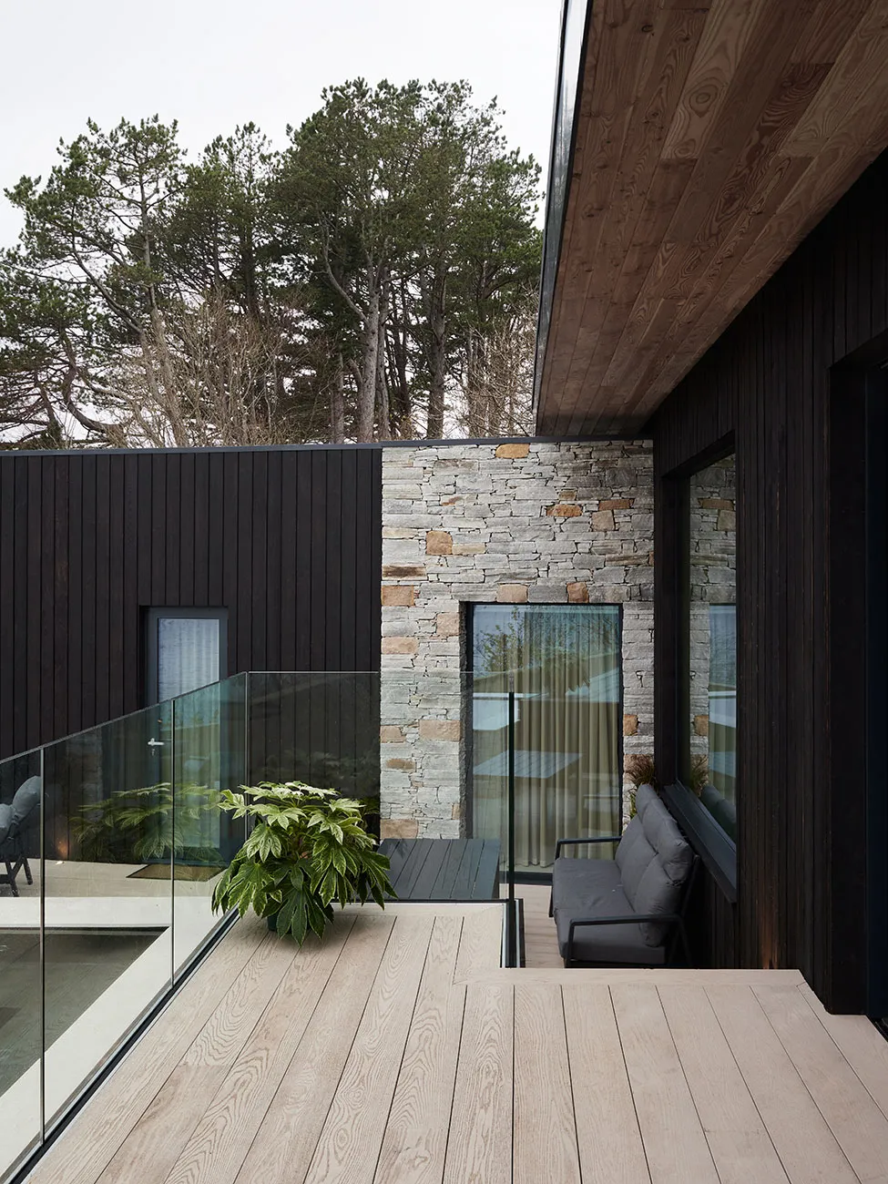 Modern outdoor living space featuring a wooden deck with a glass railing and a stone wall, surrounded by trees.