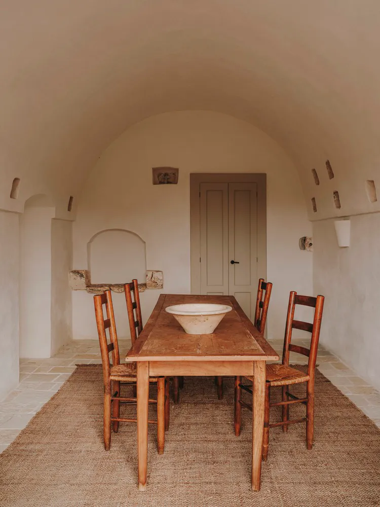 A minimalist dining room featuring a wooden table with a large bowl on it, surrounded by wooden chairs.