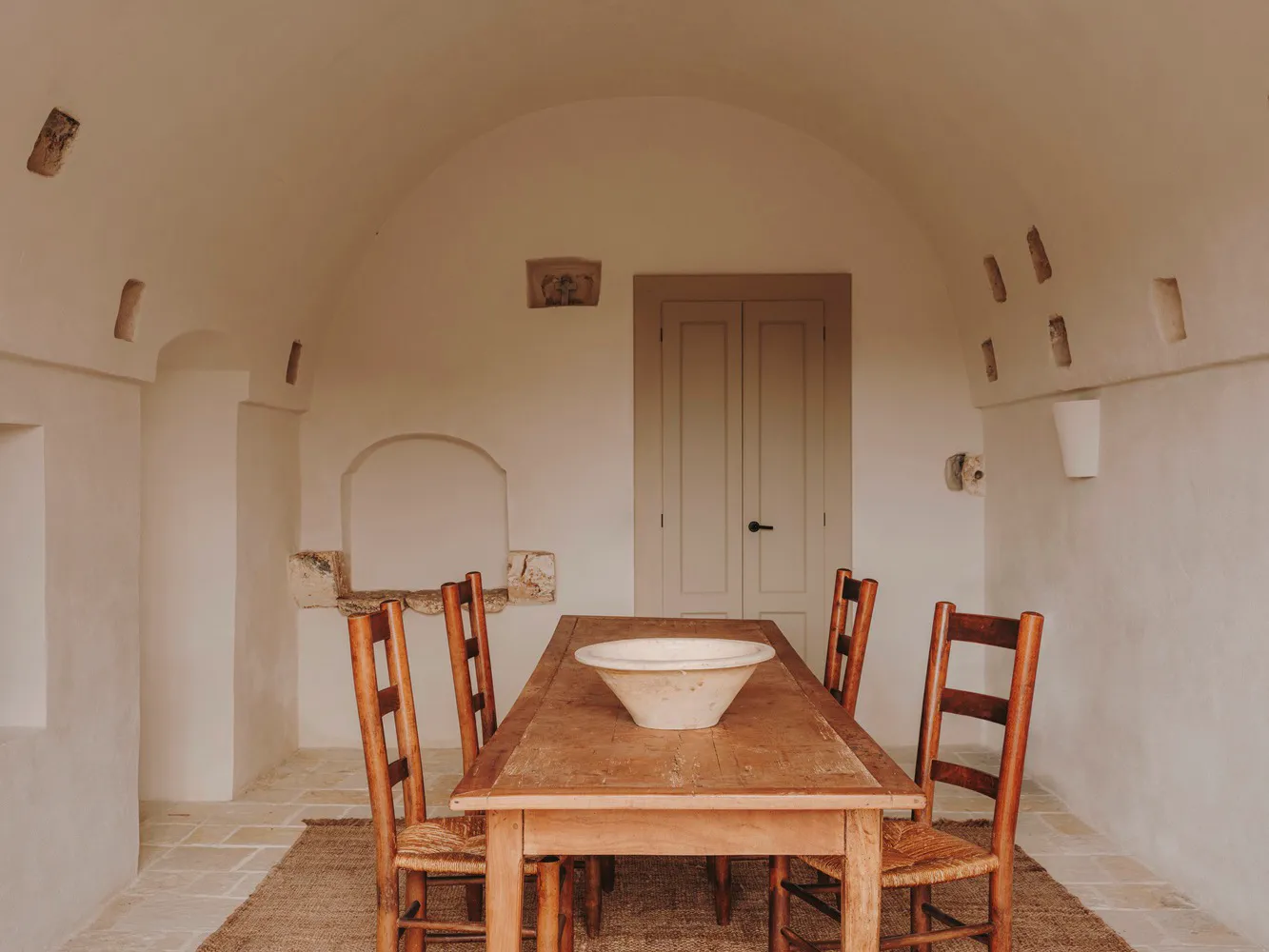 A rustic dining room with a wooden table, chairs, and a central bowl on the table. The walls are made of drywall with visible
