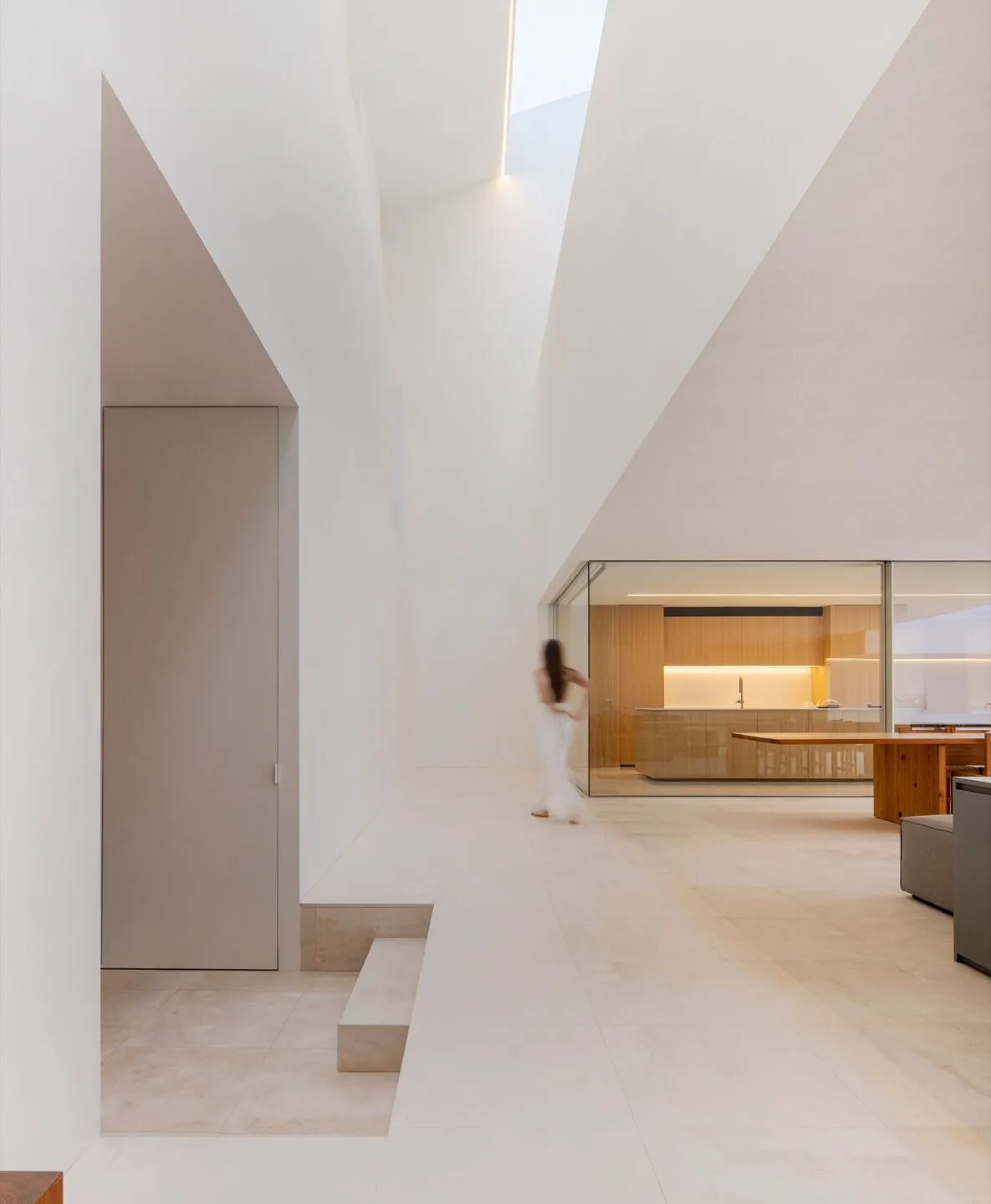 A minimalist interior space with concrete walls and floor, featuring modern furnishings and a glass-enclosed kitchen area.