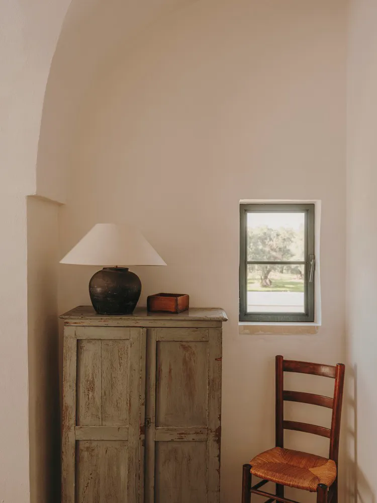 A minimalist living room featuring a wooden cabinet with a lamp and a small wooden box, positioned next to a window with a vi