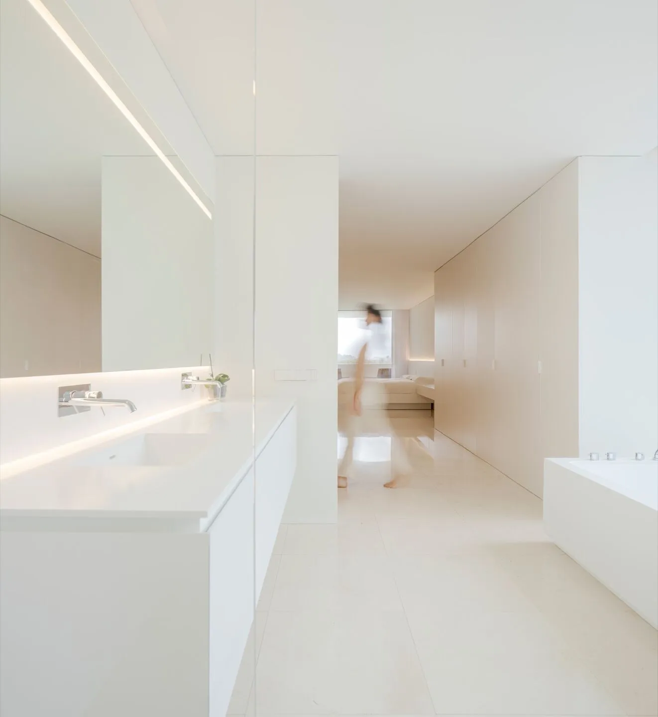 Modern minimalist bathroom with white marble flooring, fixtures, and a large mirror with lighting along the back edge.