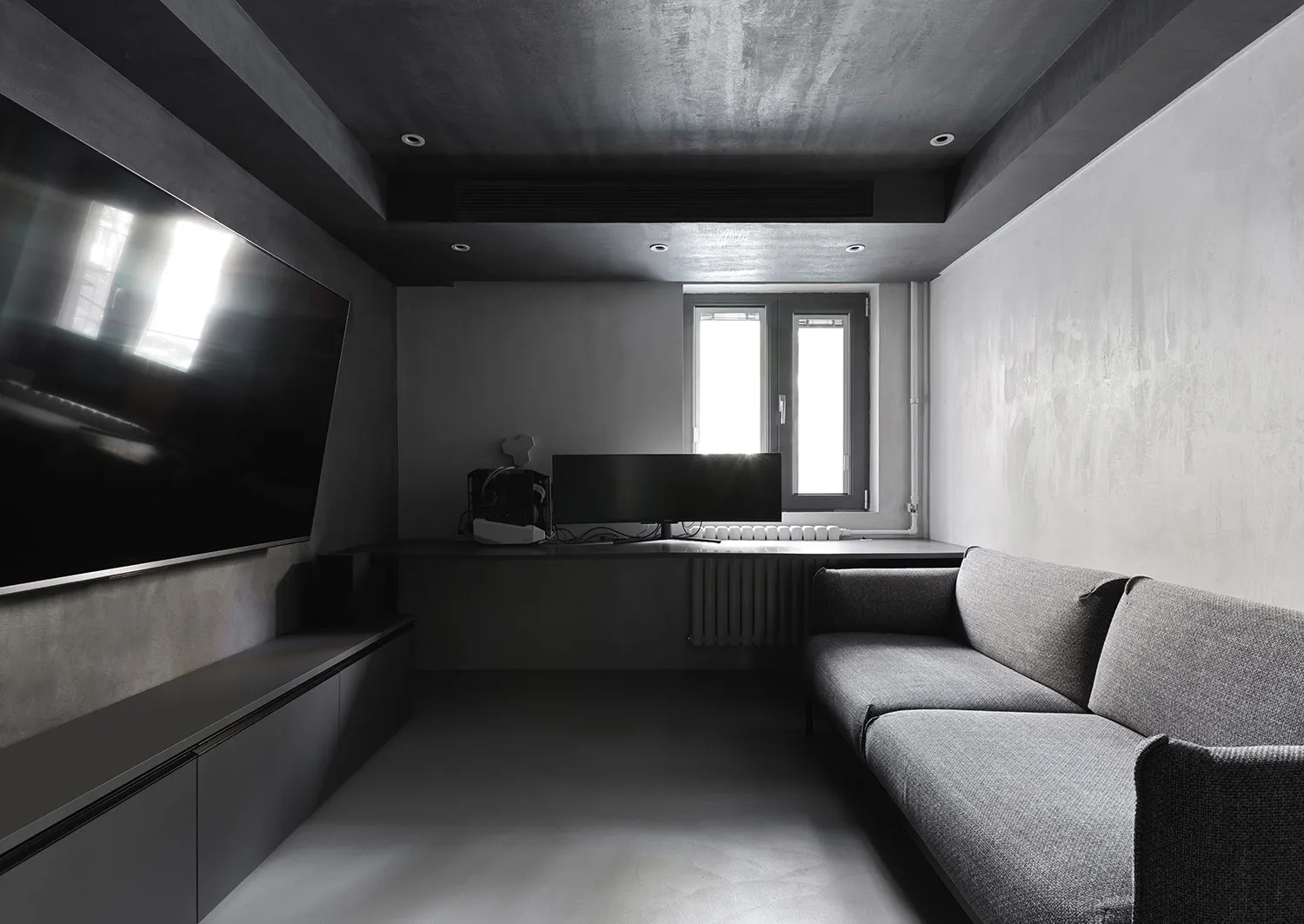 An industrial-style living room with concrete walls and floor, featuring a grey sofa and a large TV on the left wall.