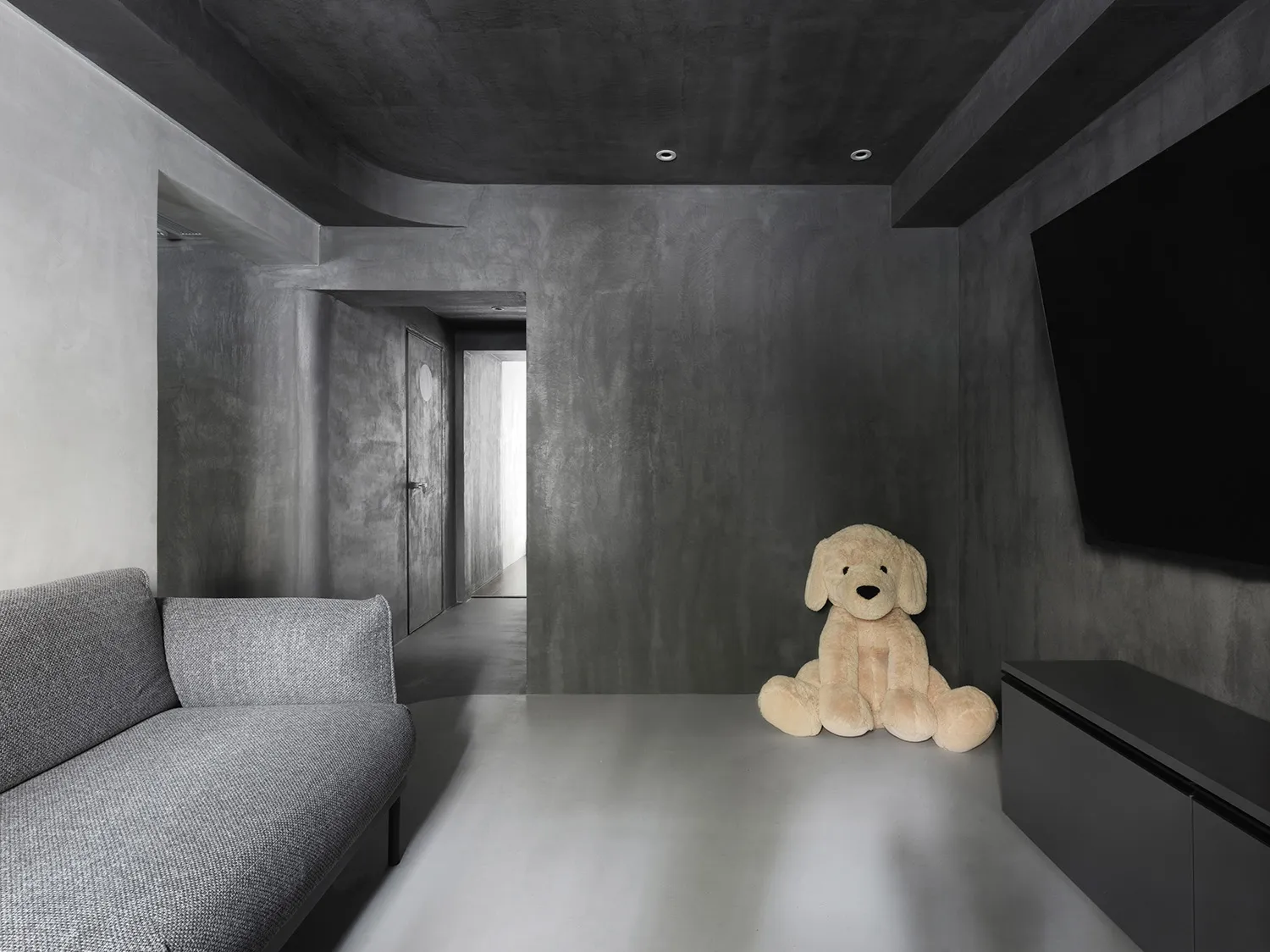 A minimalist living room with a gray sofa, a large stuffed dog, and a flat-screen TV mounted on the wall, featuring textured