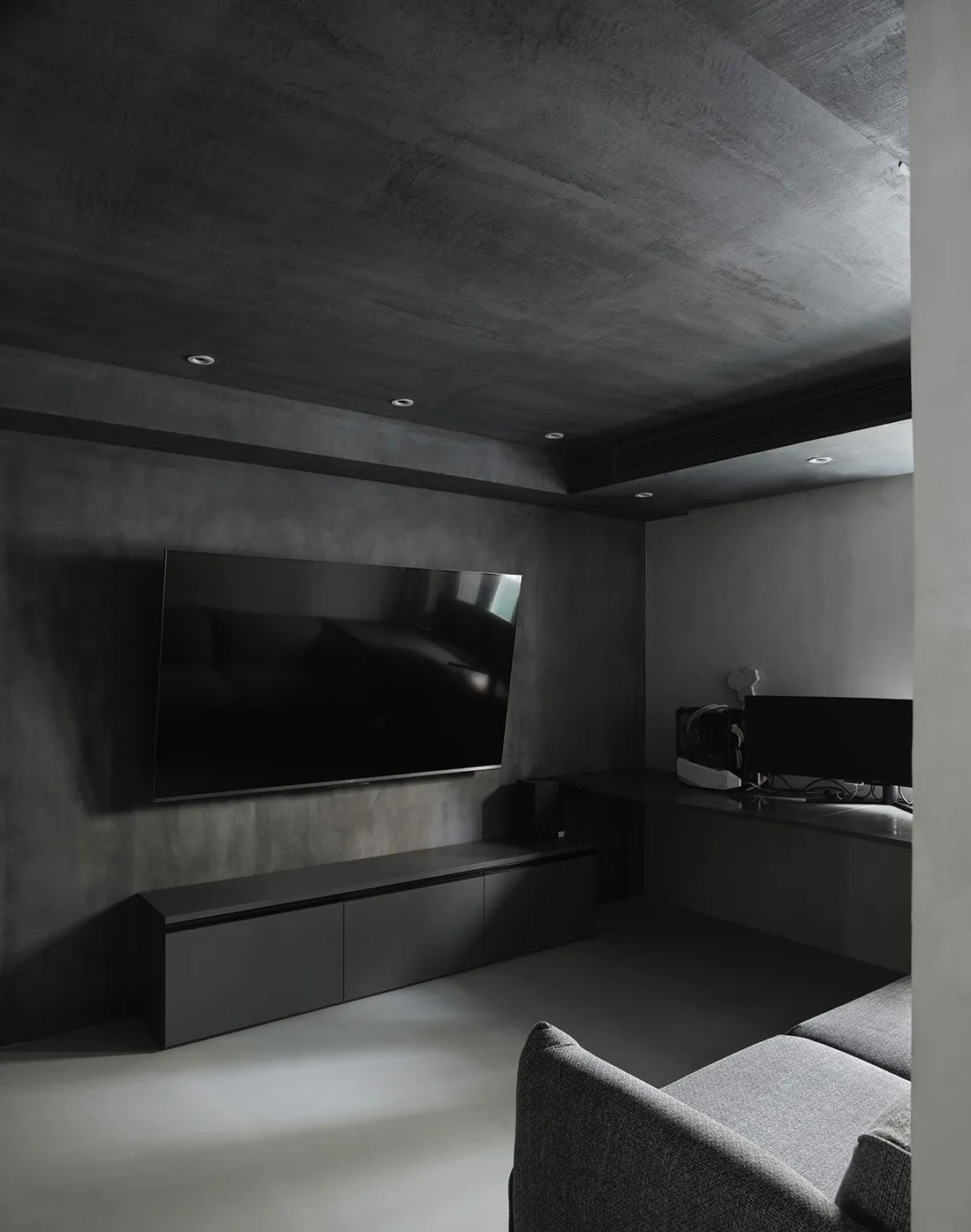 Modern industrial-style living room with concrete walls and floor, featuring a flat-screen TV and grey upholstered furniture.