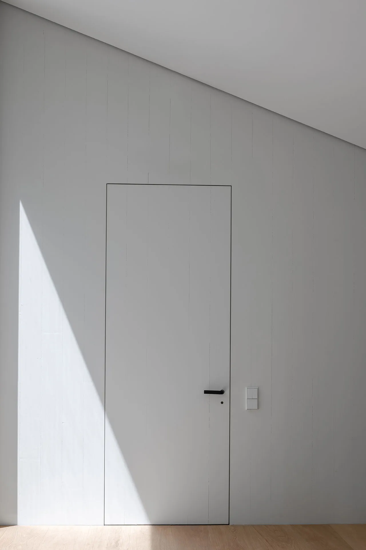 A minimalist white door with a light shadow cast on it, set against a white wall with vertical paneling.