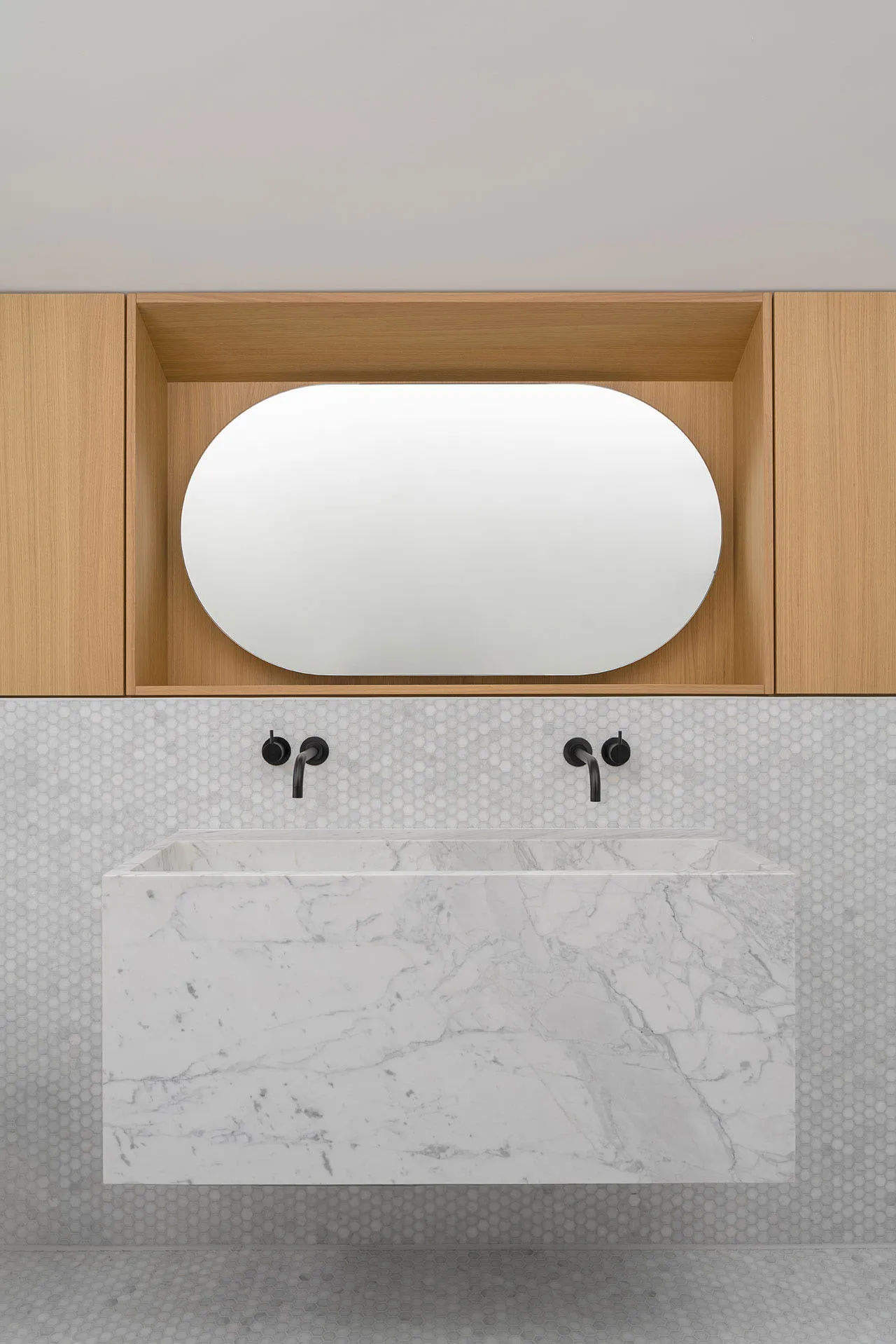 A minimalist bathroom with white marble floating vanity, black faucets, white hexagonal tiles, and light wood cabinets.