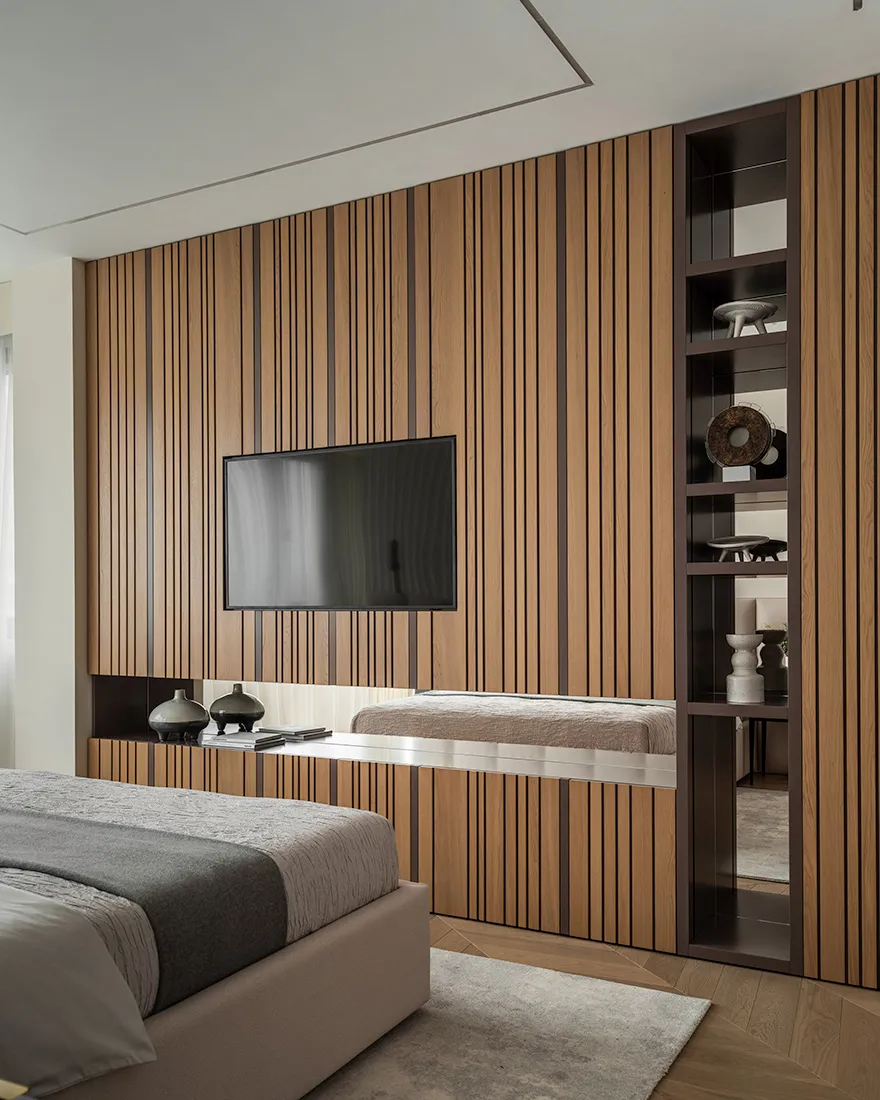 A minimalist bedroom with a brown paneled wall, a white bed with a gray blanket, and a floor-to-ceiling shelf with decorative