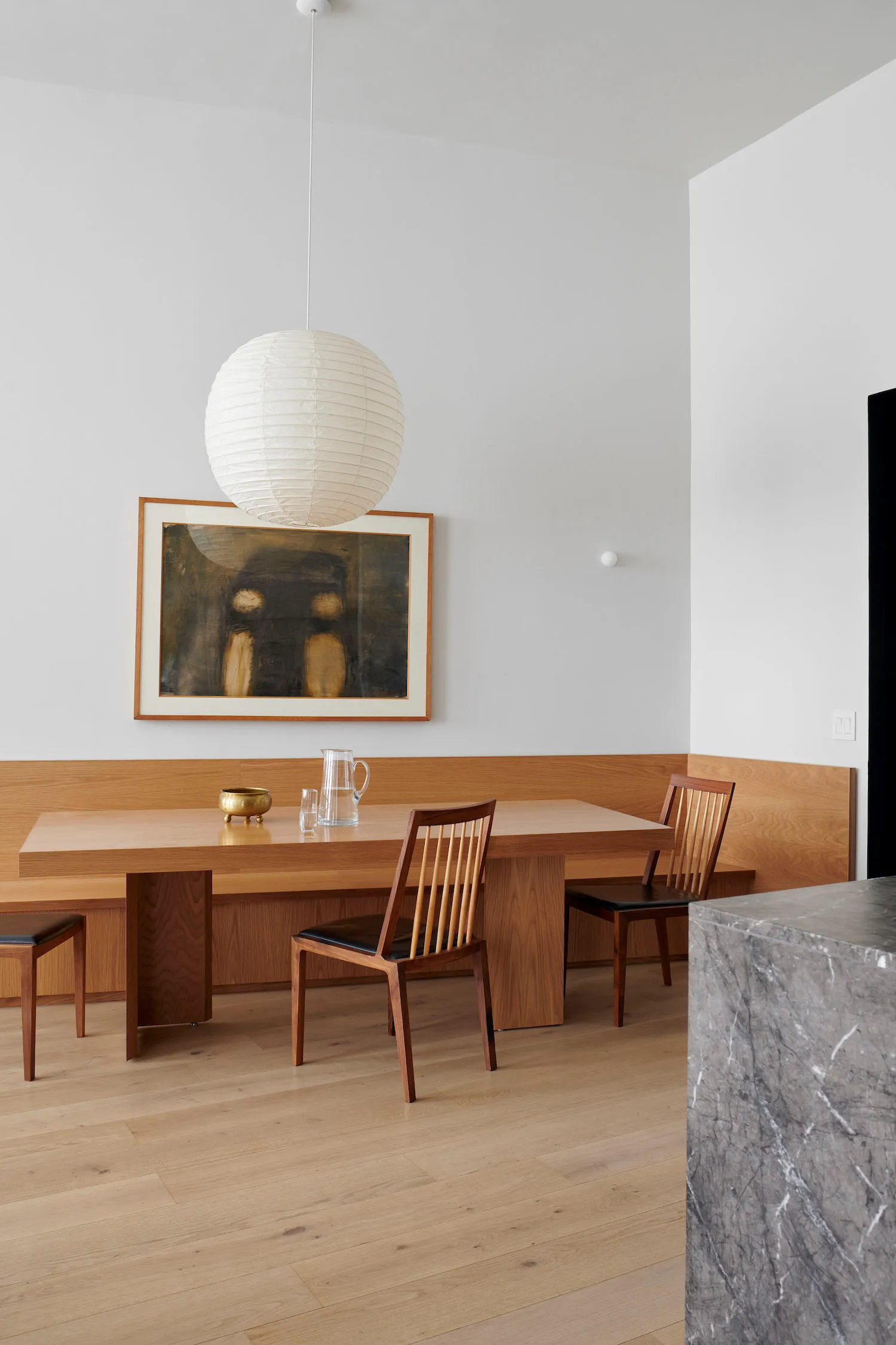 Modern minimalist dining area with a wooden bench and chairs, white pendant light fixture, and abstract painting on the wall.