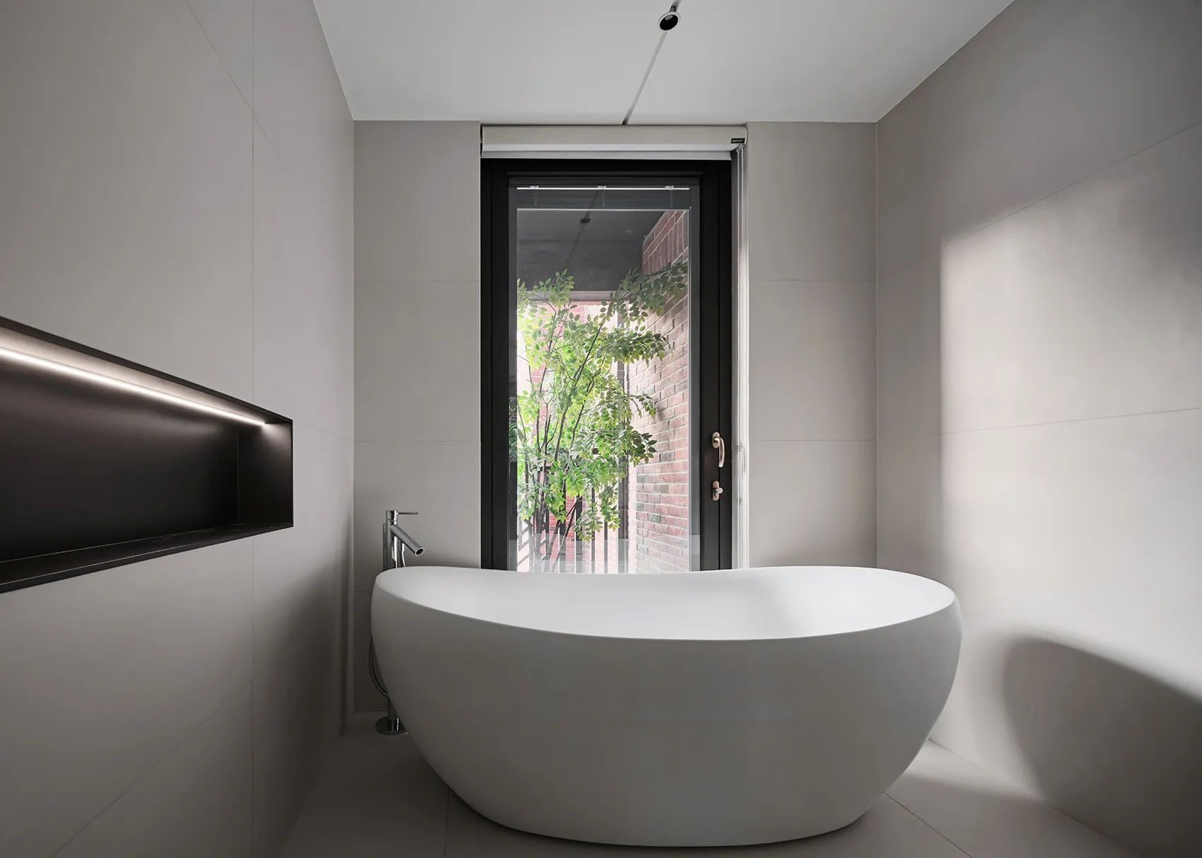 A modern minimalist bathroom with a white porcelain bathtub, grey porcelain tiles, and a large window with a view of greenery