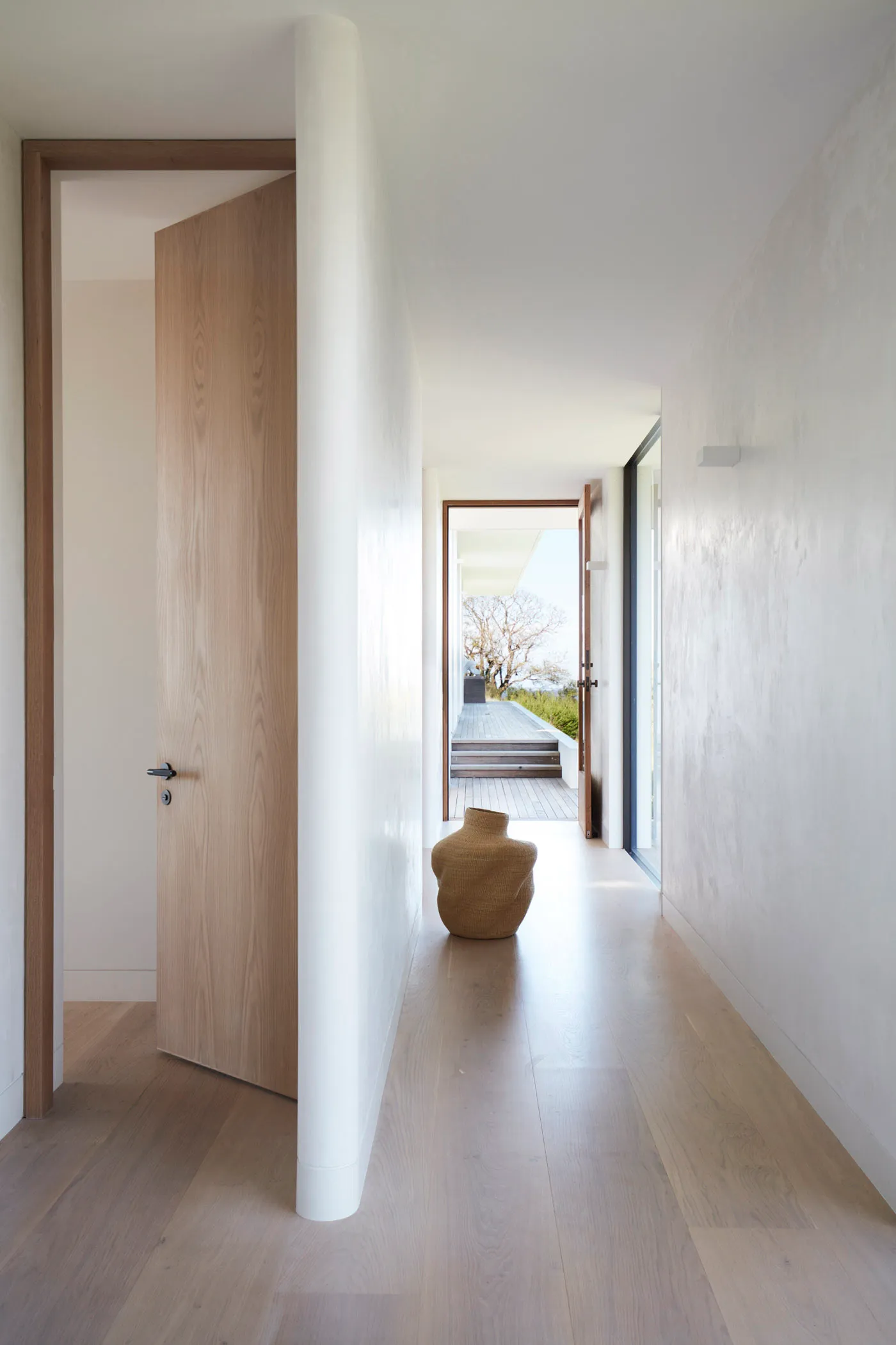 A modern interior hallway with wooden flooring and walls, featuring an open door and a vase on the floor.