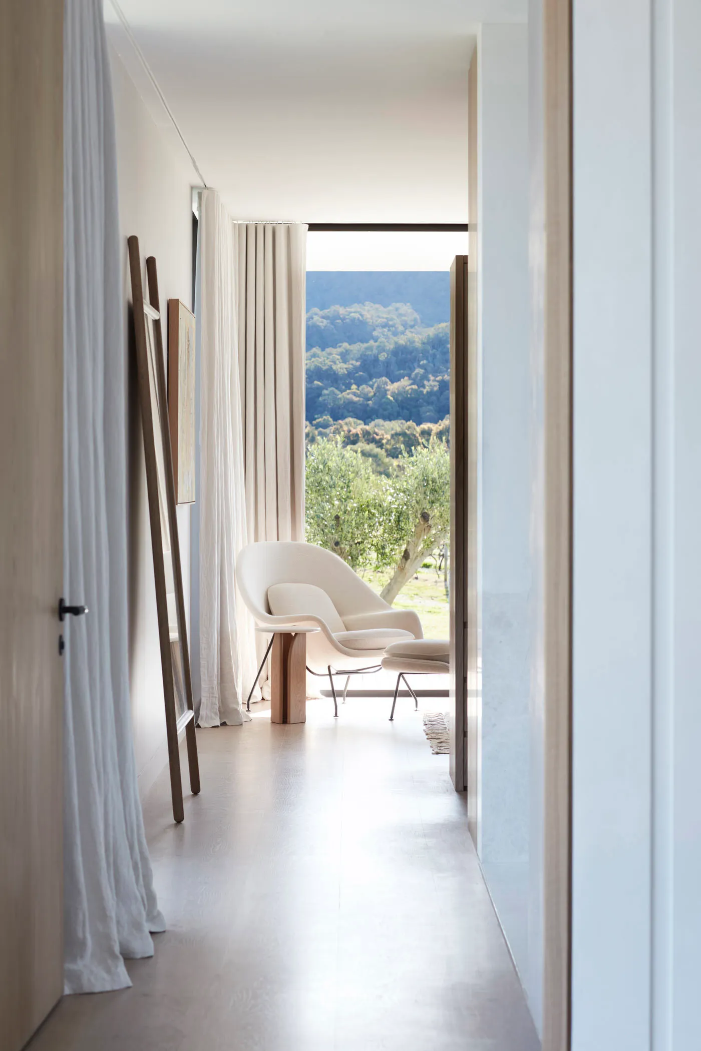 Minimalist modern living room with white furniture and wooden elements, featuring a view of greenery outside through the larg