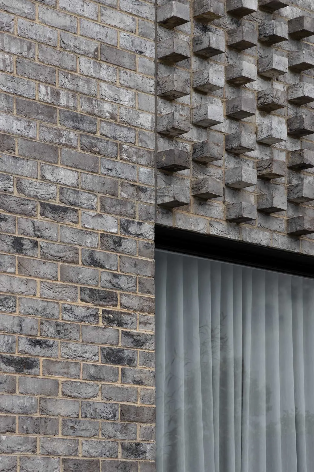 Grey brick wall with protruding elements and a window with sheer curtains.