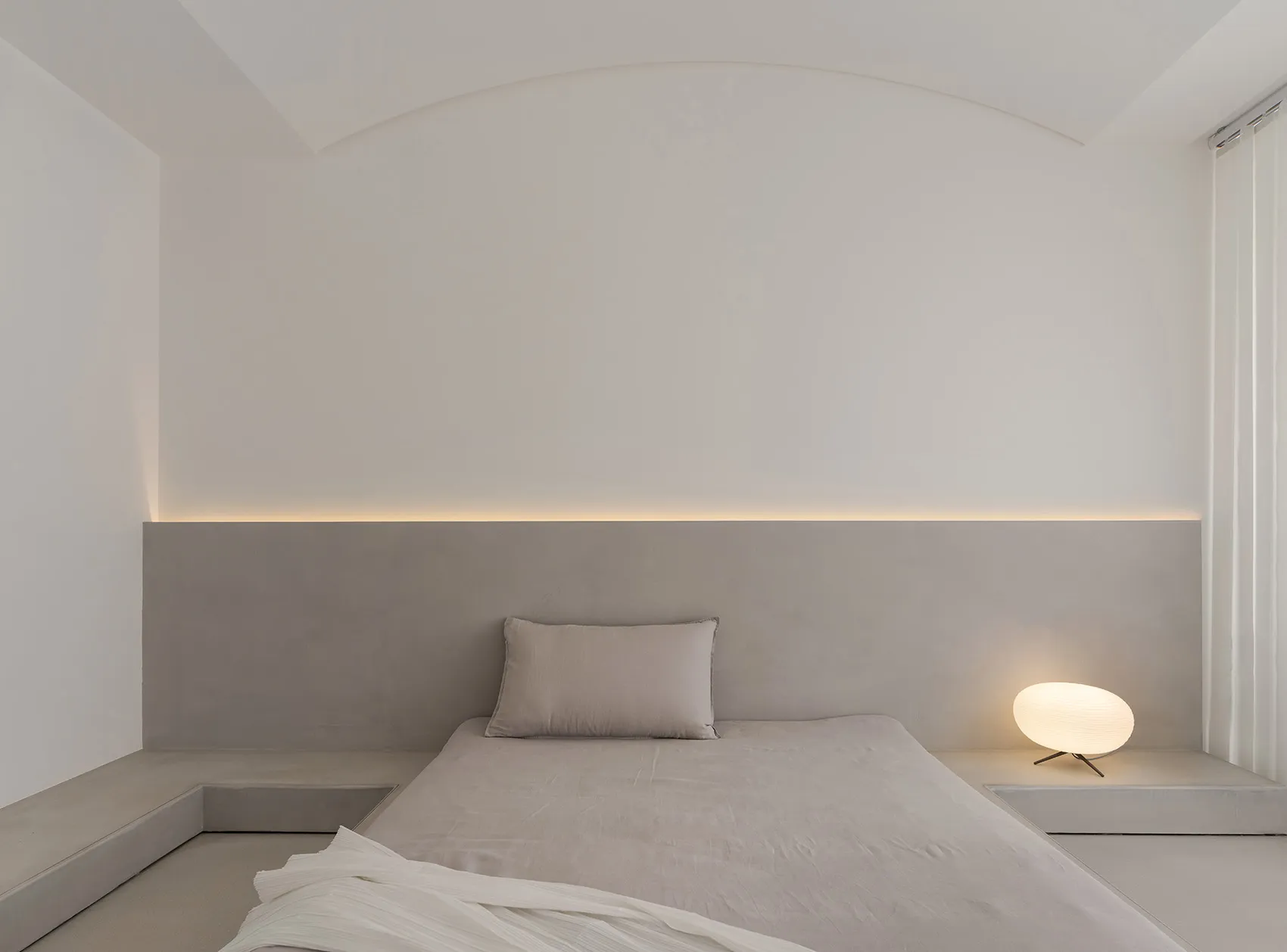 Minimalist bedroom with concrete walls and floor, featuring a bed with simple bedding and a small round lamp.