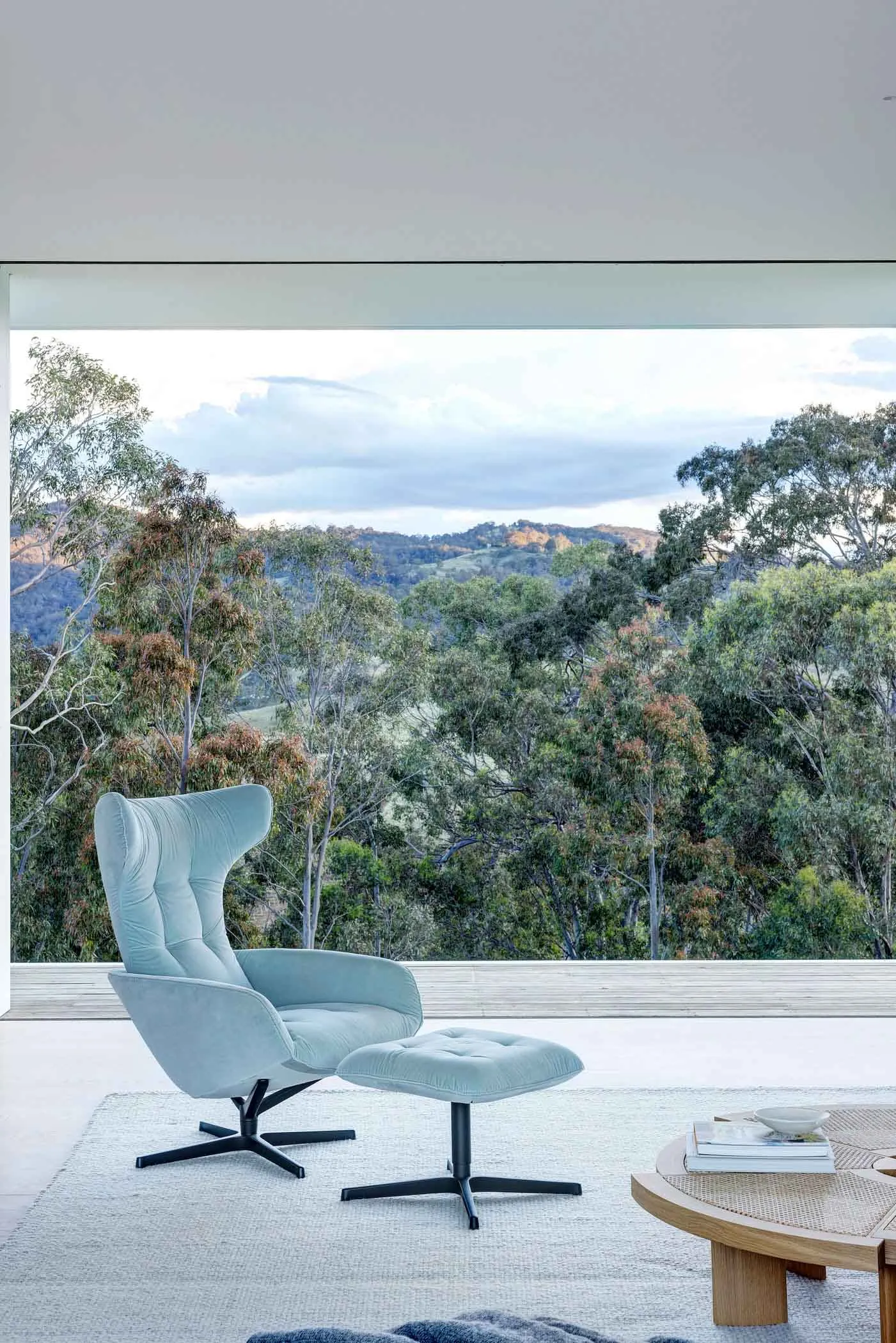 Modern living room with a blue upholstered armchair and ottoman, featuring a scenic outdoor view of trees and hills.