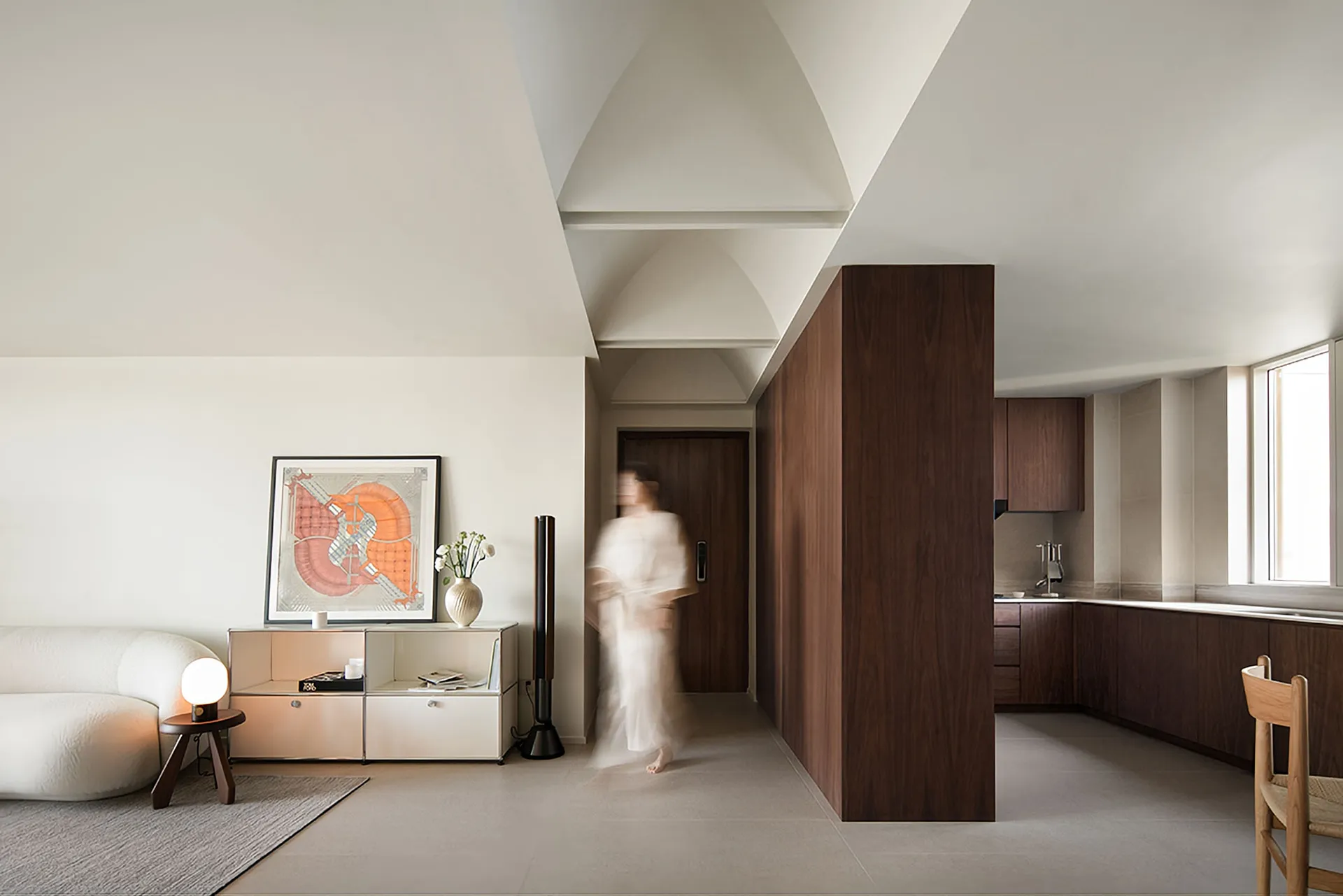 A minimalist modern living room featuring a white sofa, a wooden pillar, and a kitchen area with dark wooden cabinets.
