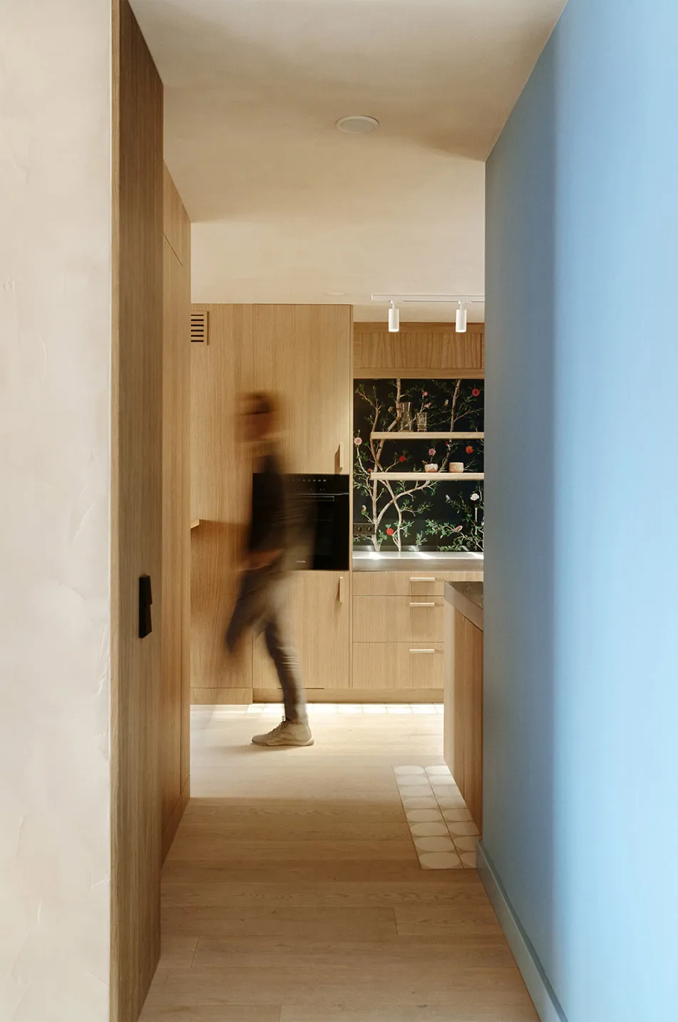 A person walking in a modern kitchen with wooden cabinets and a blue wall.