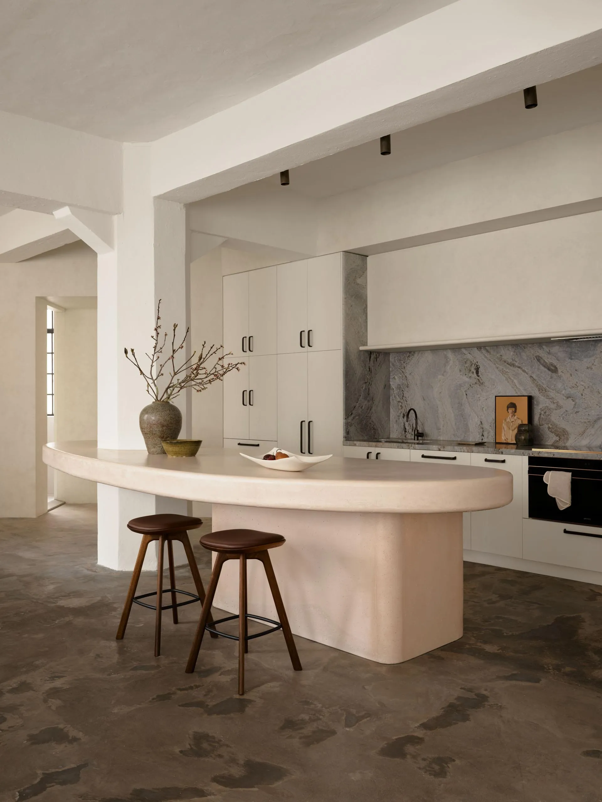 A minimalist residential kitchen featuring a concrete island with a smooth, curved countertop, two wooden stools, a vase, and