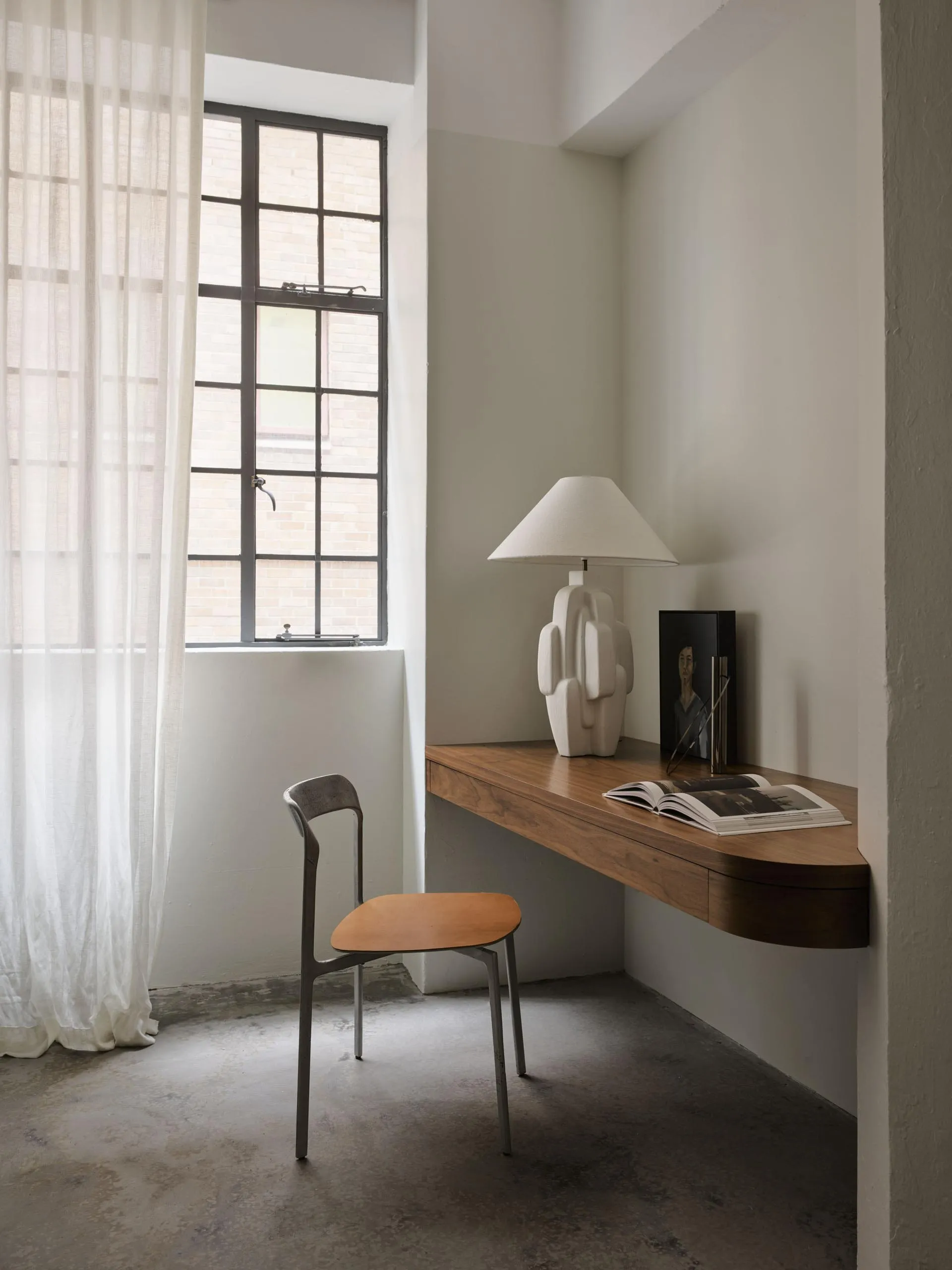 A minimalist office space featuring a wooden desk setup with a white lamp, open book, and portrait on the wall, alongside she