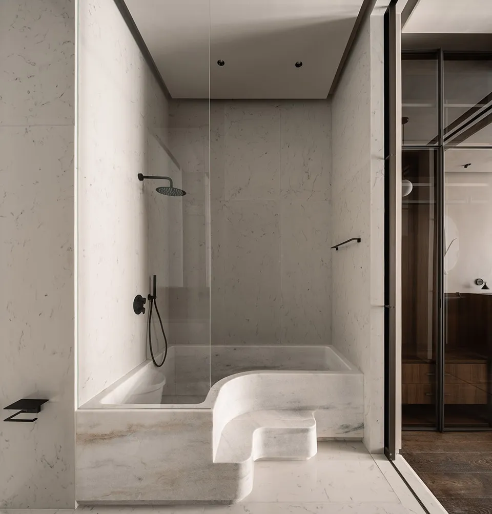 A modern minimalist bathroom with marble walls and floor, featuring a built-in bathtub with a sleek glass shower enclosure an