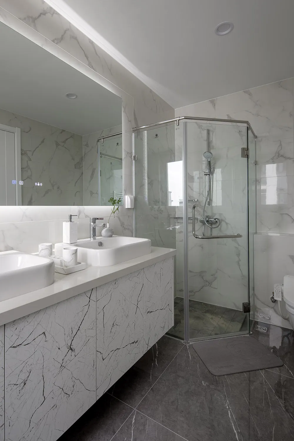 A modern bathroom with marble walls and floor, featuring a glass-enclosed shower, a floating vanity with white ceramic sinks,