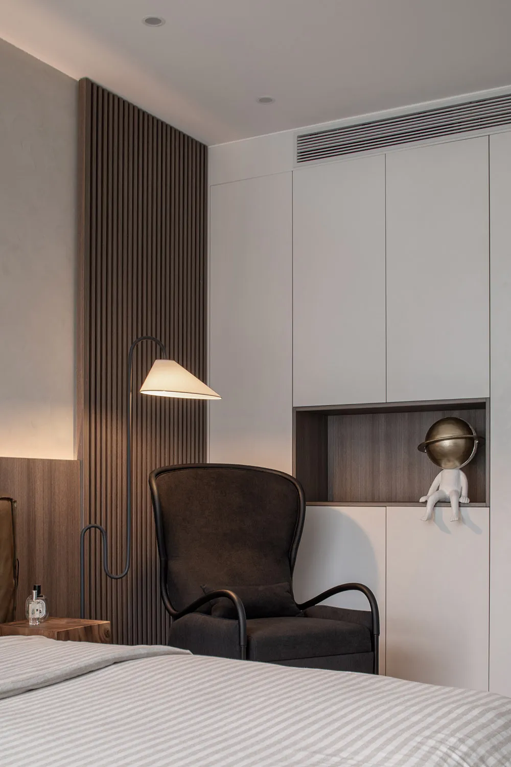Modern bedroom with a dark wooden accent wall, a dark armchair, a floor lamp, and a white bed with striped sheets.