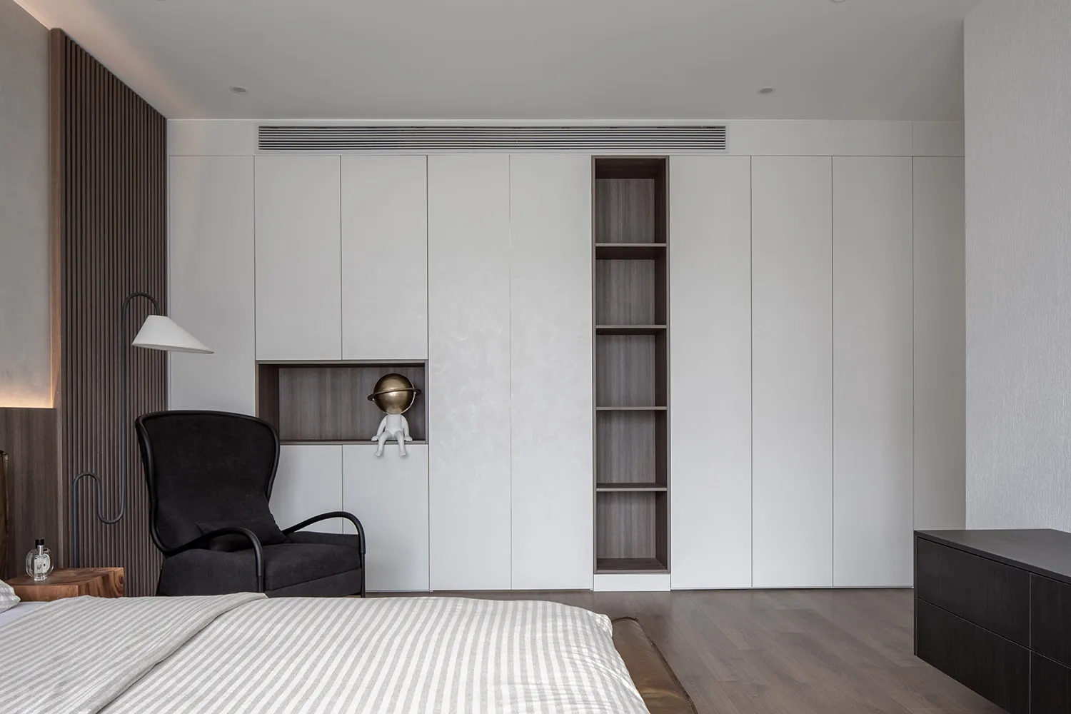 A modern bedroom with white paneled walls, wooden pine floors, and a black armchair with a lamp.