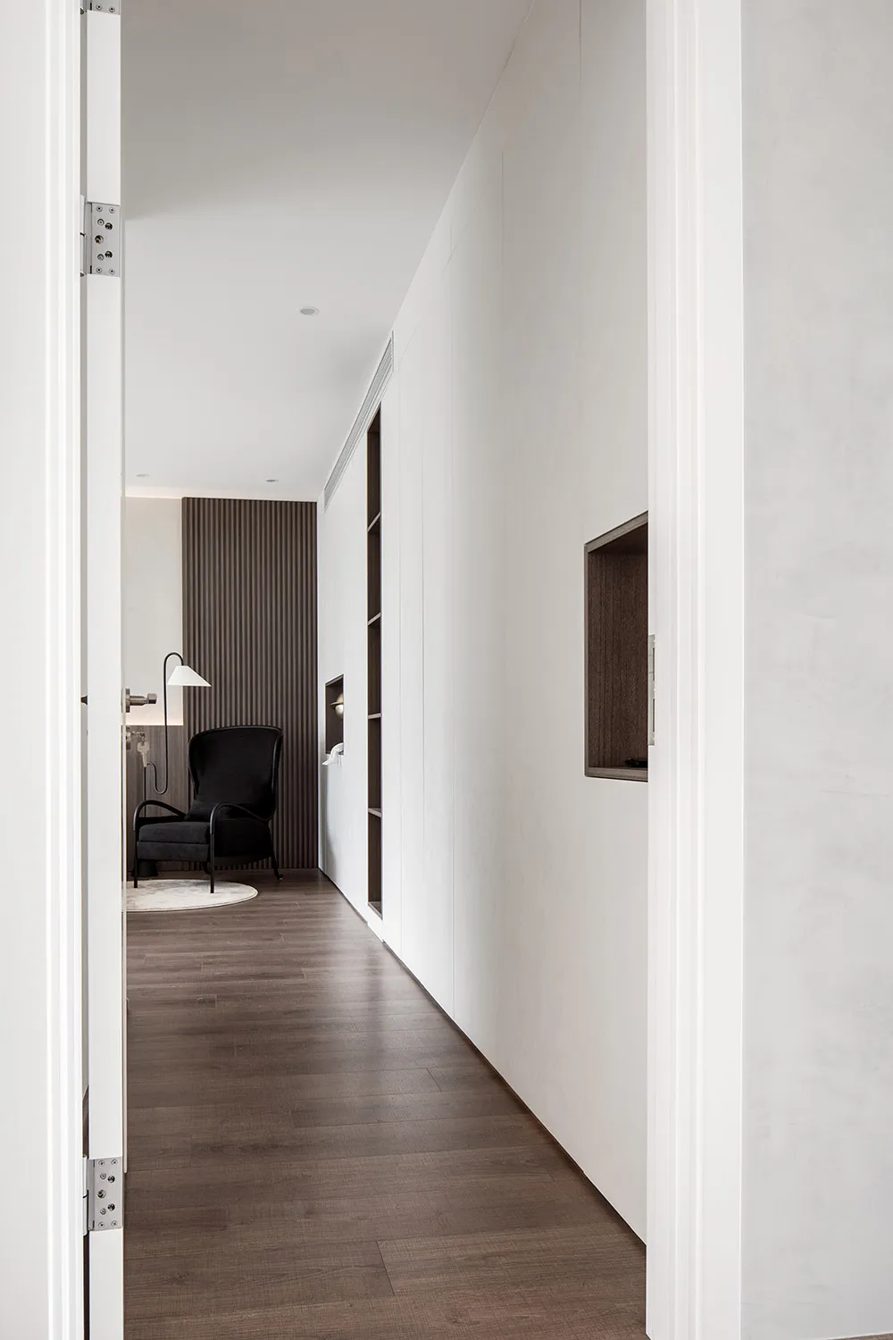 Minimalist white corridor with wooden flooring, drywall walls, and a black armchair at the end.