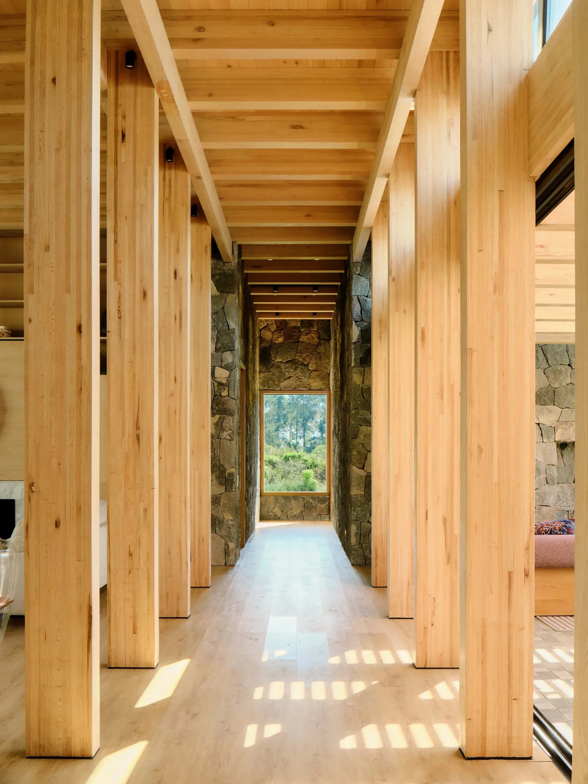 A modern corridor with wooden columns and a stone wall, featuring a window overlooking a natural landscape.