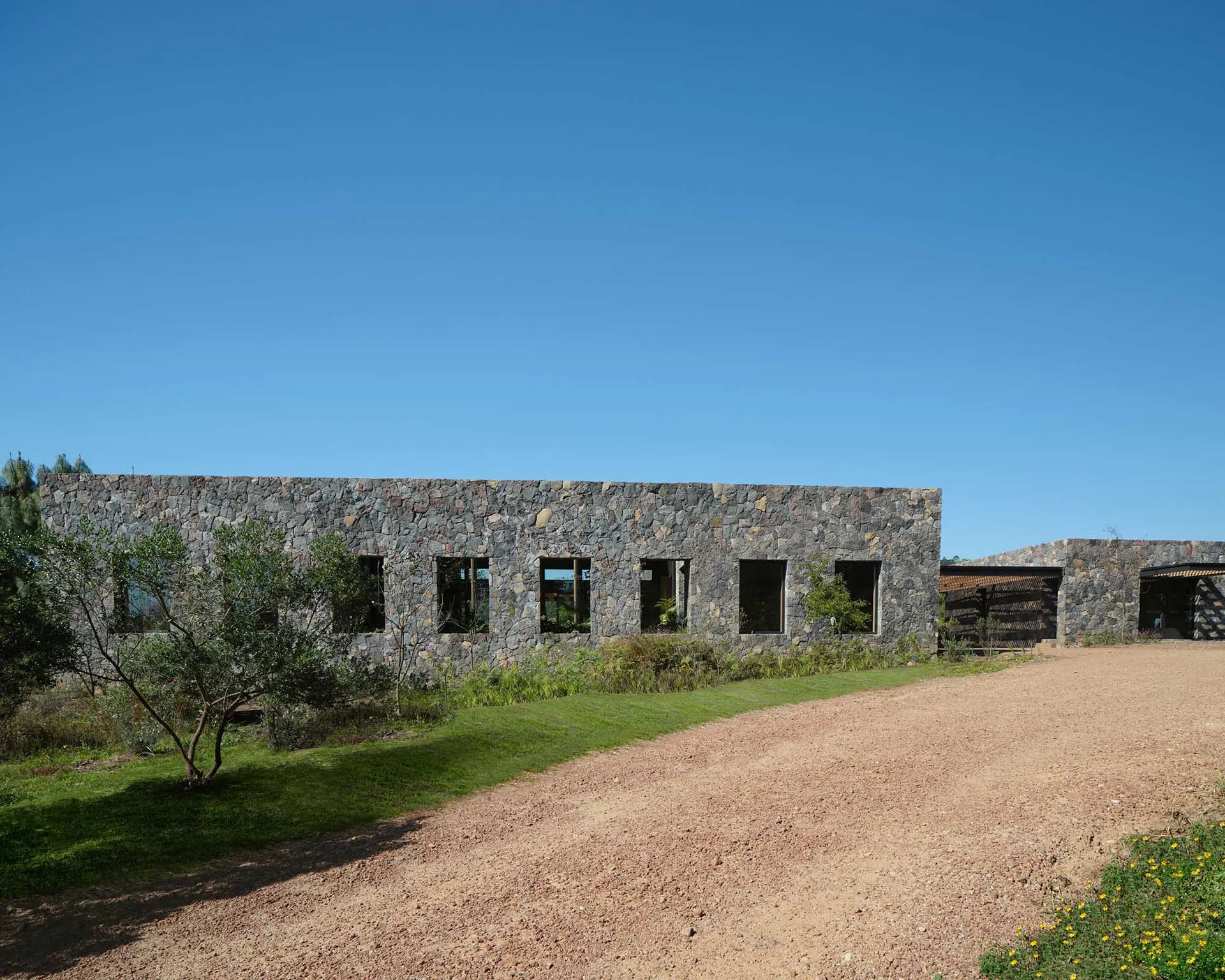 A modern building constructed of stone with a dirt driveway and greenery.