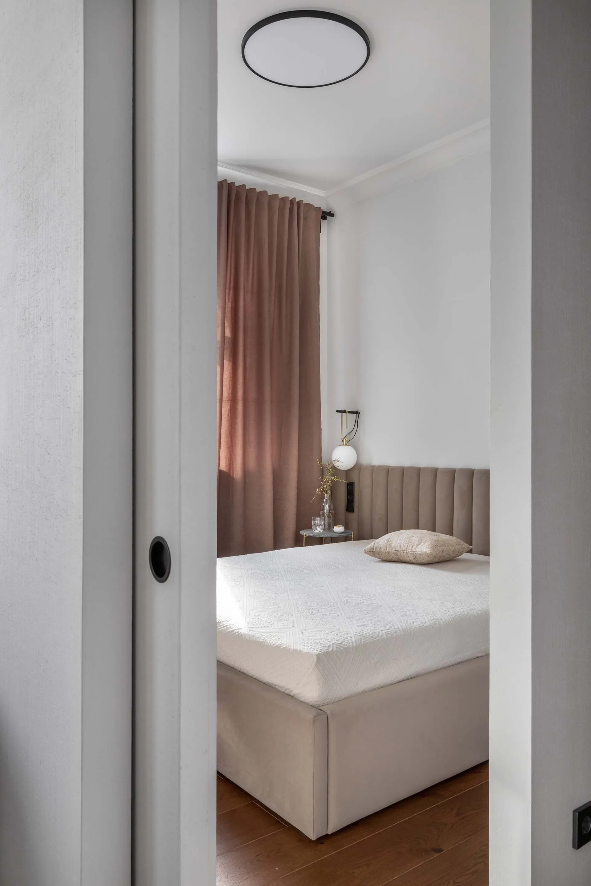 A minimalist bedroom with a beige upholstered bed, brown velvet curtains, and wooden floor.