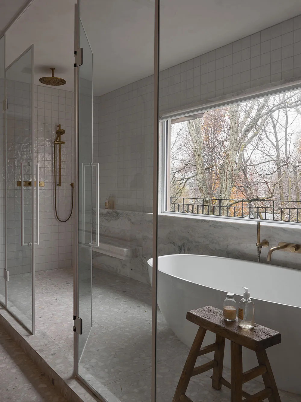 Modern minimalist bathroom featuring a white marble tub, glass shower enclosure, and a window overlooking trees.