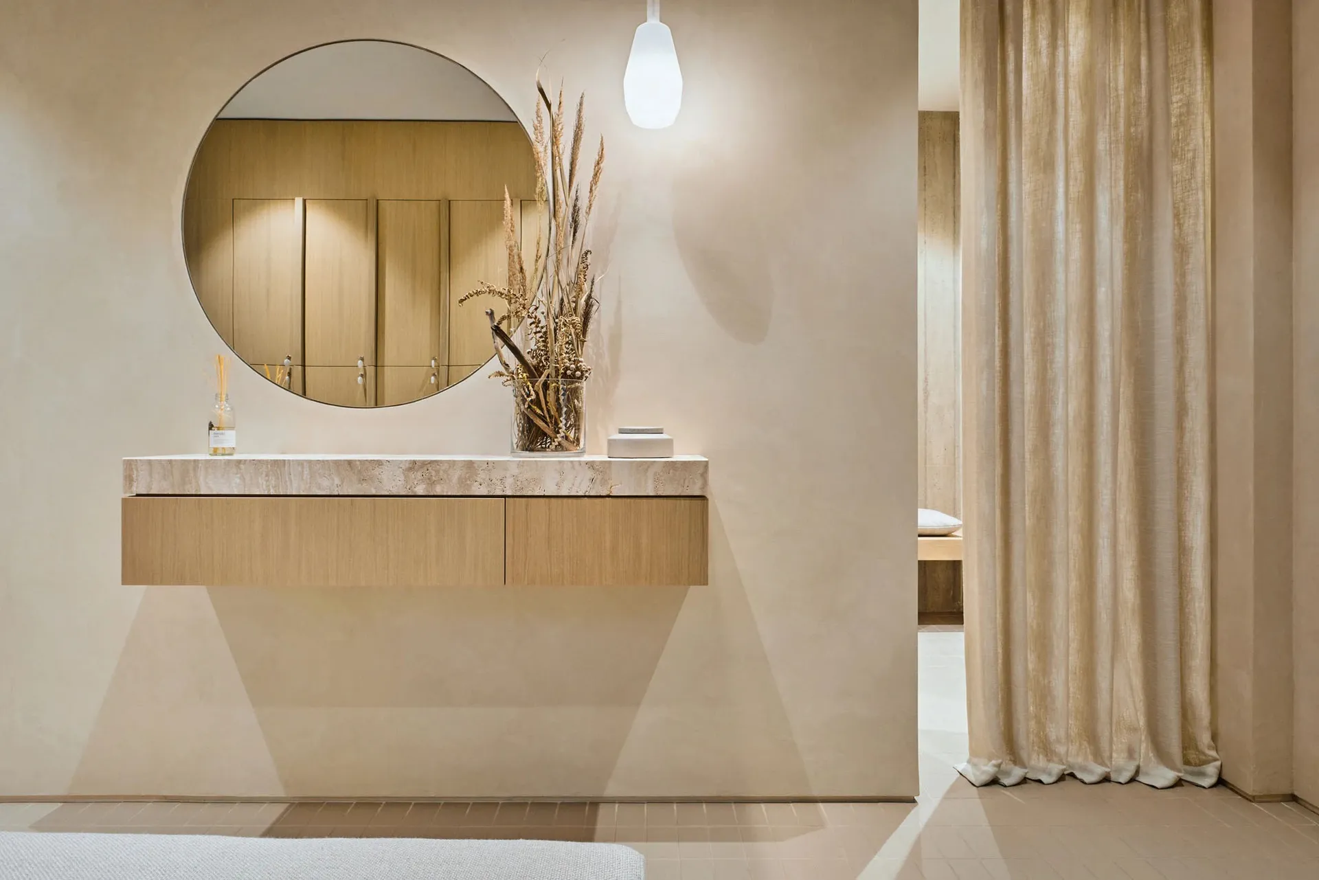 A modern bathroom featuring a marble countertop with a vase of dried plants and a round mirror above it.