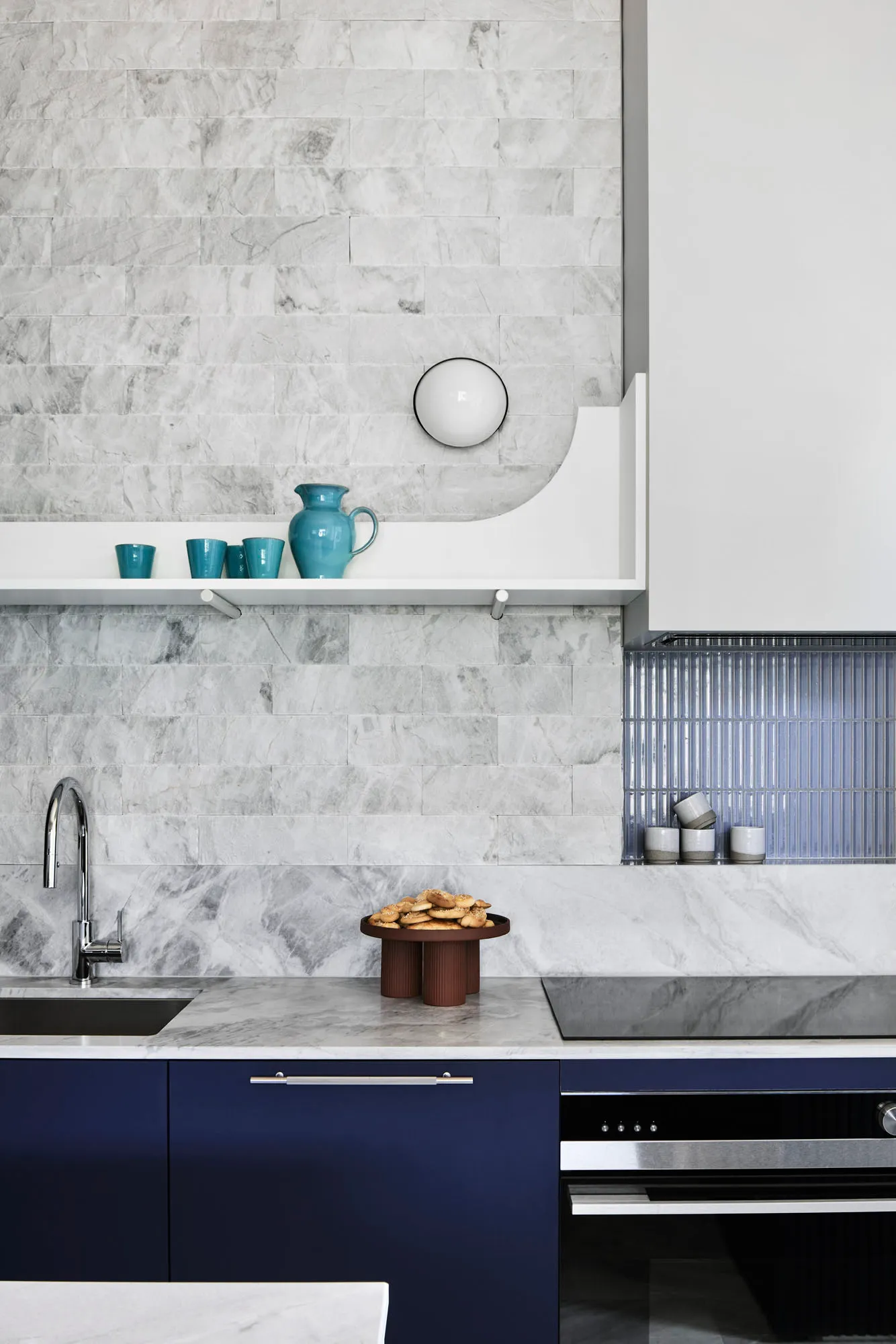 A modern kitchen with marble countertops and a blue cabinet beneath, featuring a marble backsplash, blue cups on a shelf, and