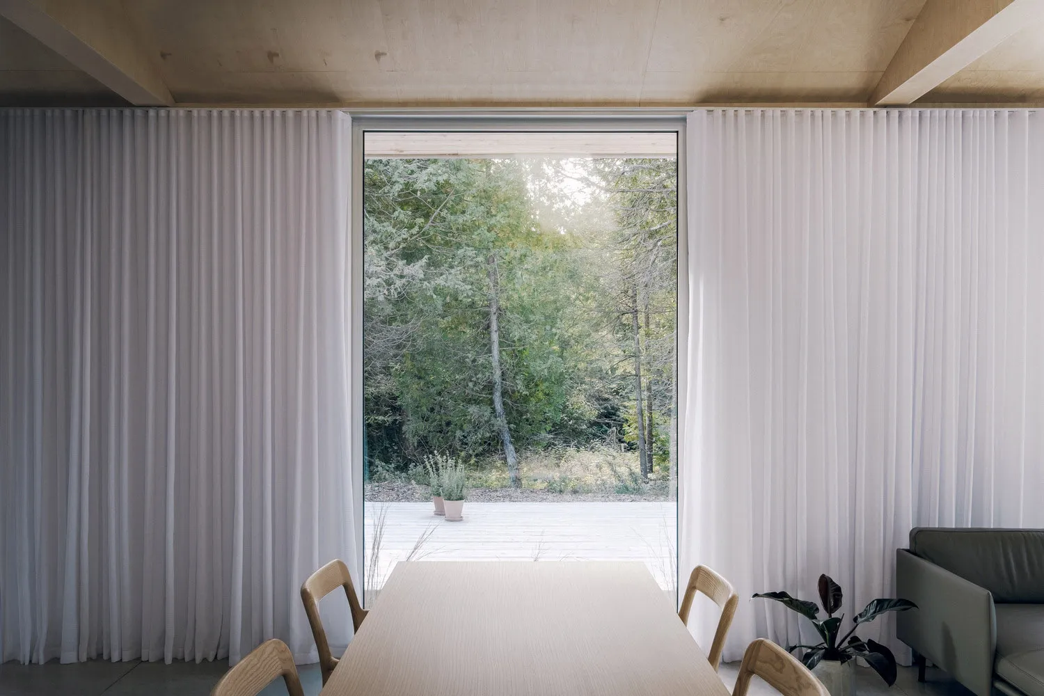 A minimalist dining room featuring white curtains and wooden furniture, offering a serene view of the forest through a large