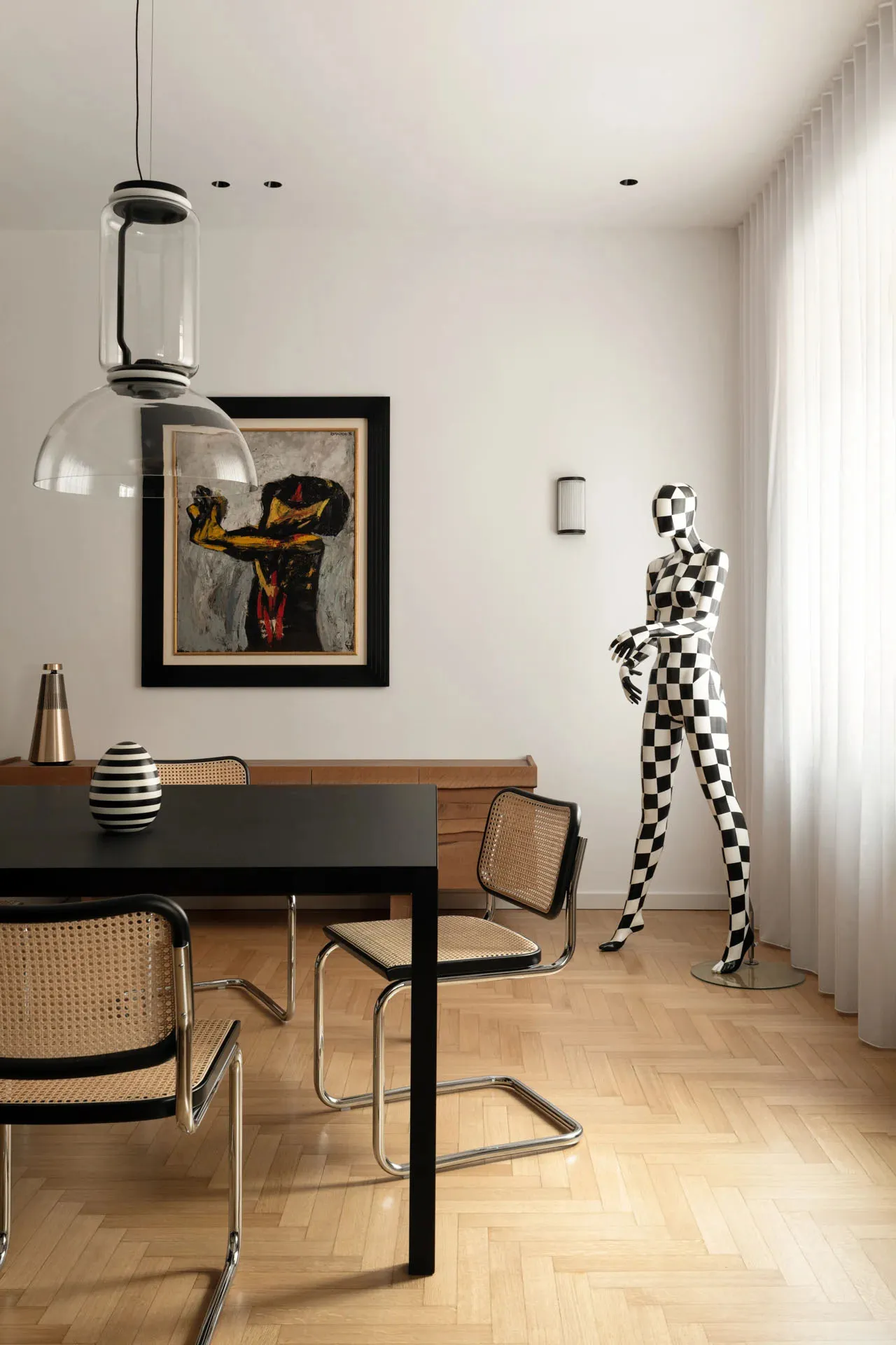 A minimalist dining room featuring a black table with woven cane chairs, herringbone wooden flooring, and a black and white c