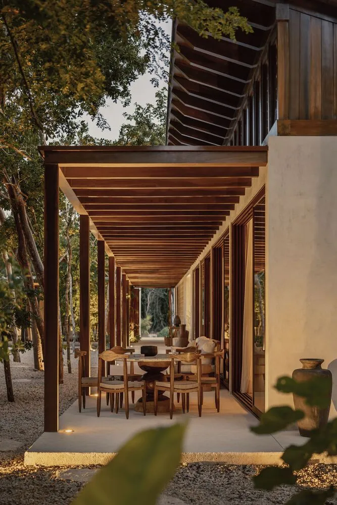 A wooden patio structure with furniture underneath, surrounded by trees and gravel.