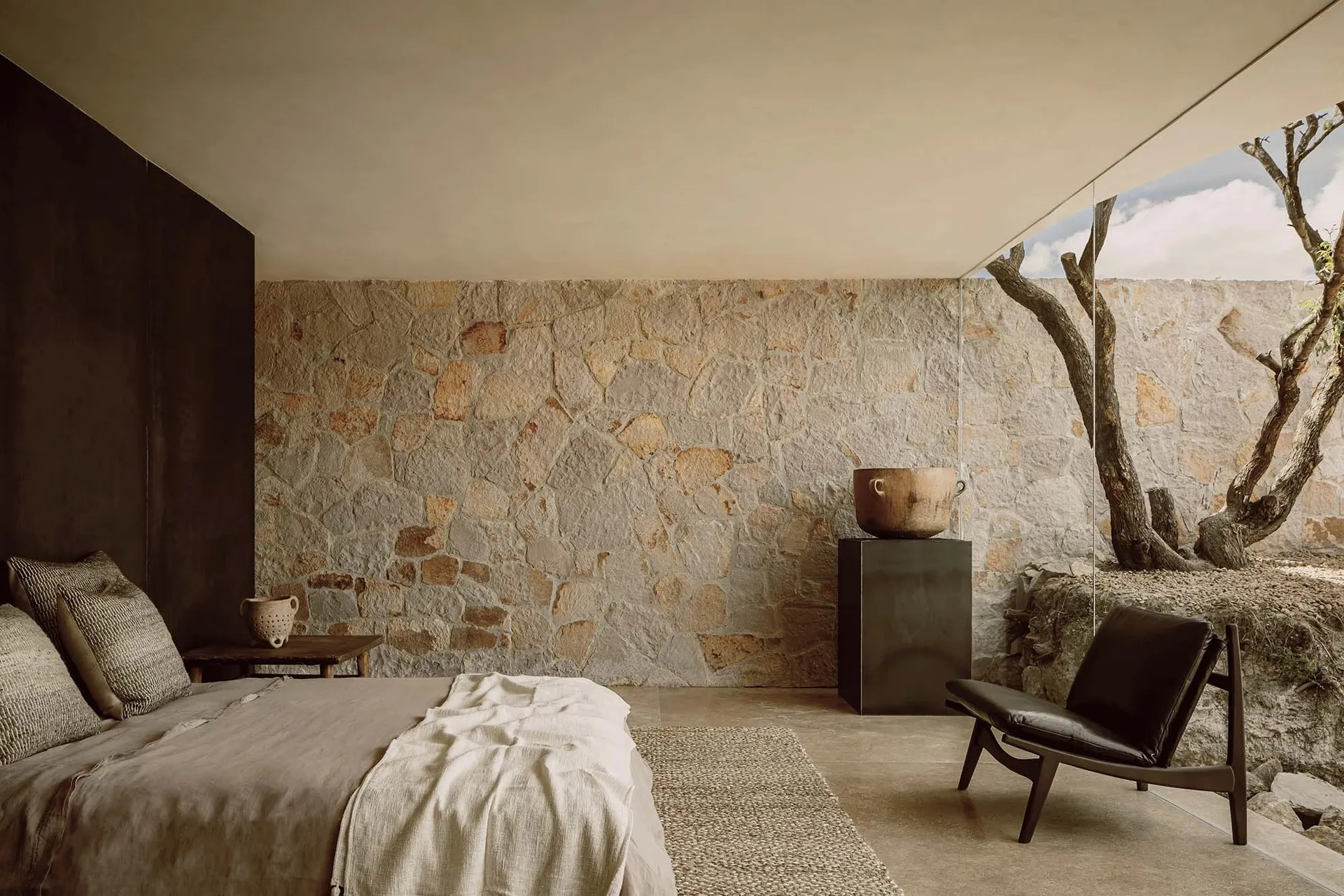 A minimalist bedroom featuring a stone wall, concrete floor, and natural surroundings with trees.