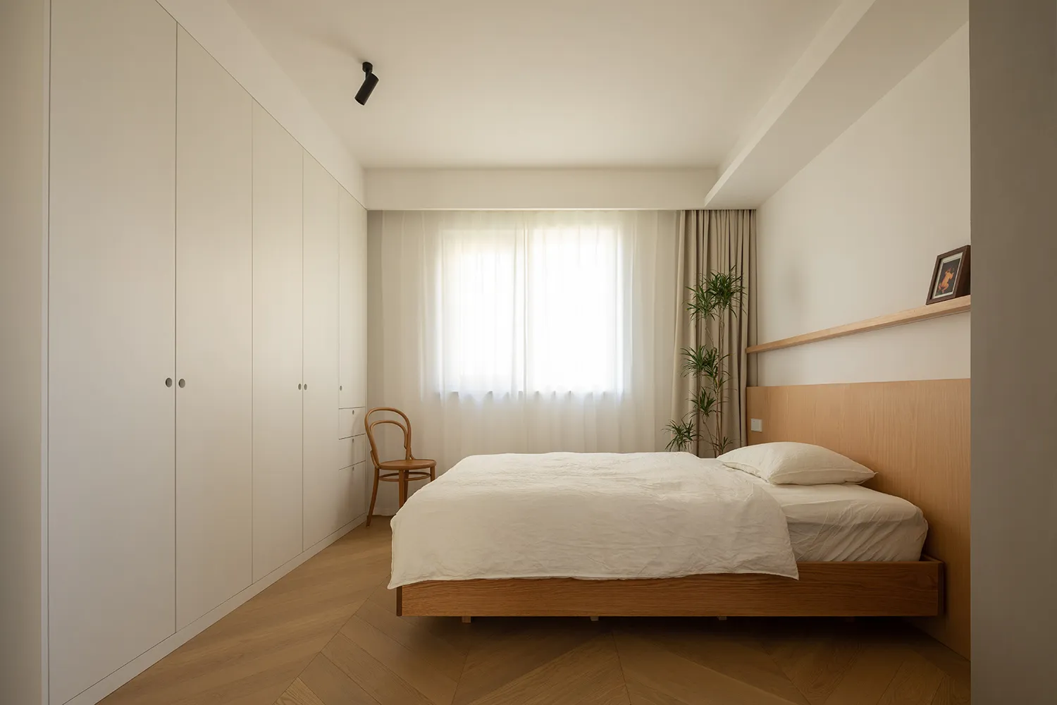 A modern minimalist bedroom featuring a wooden bed and furniture, with a large window and a white wall.
