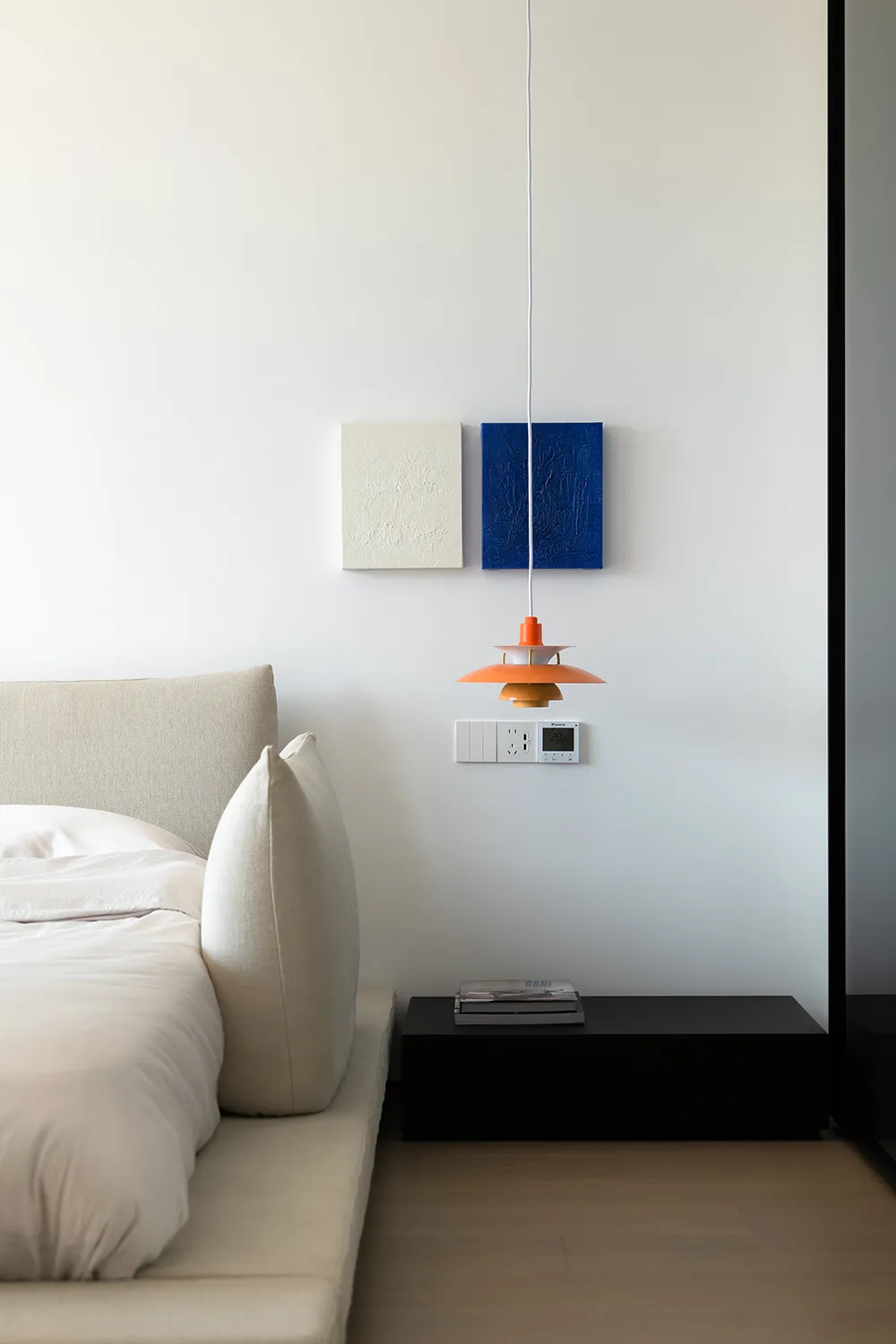 A minimalist bedroom with a bed, pillows, and a hanging lamp against a white wall with framed art.