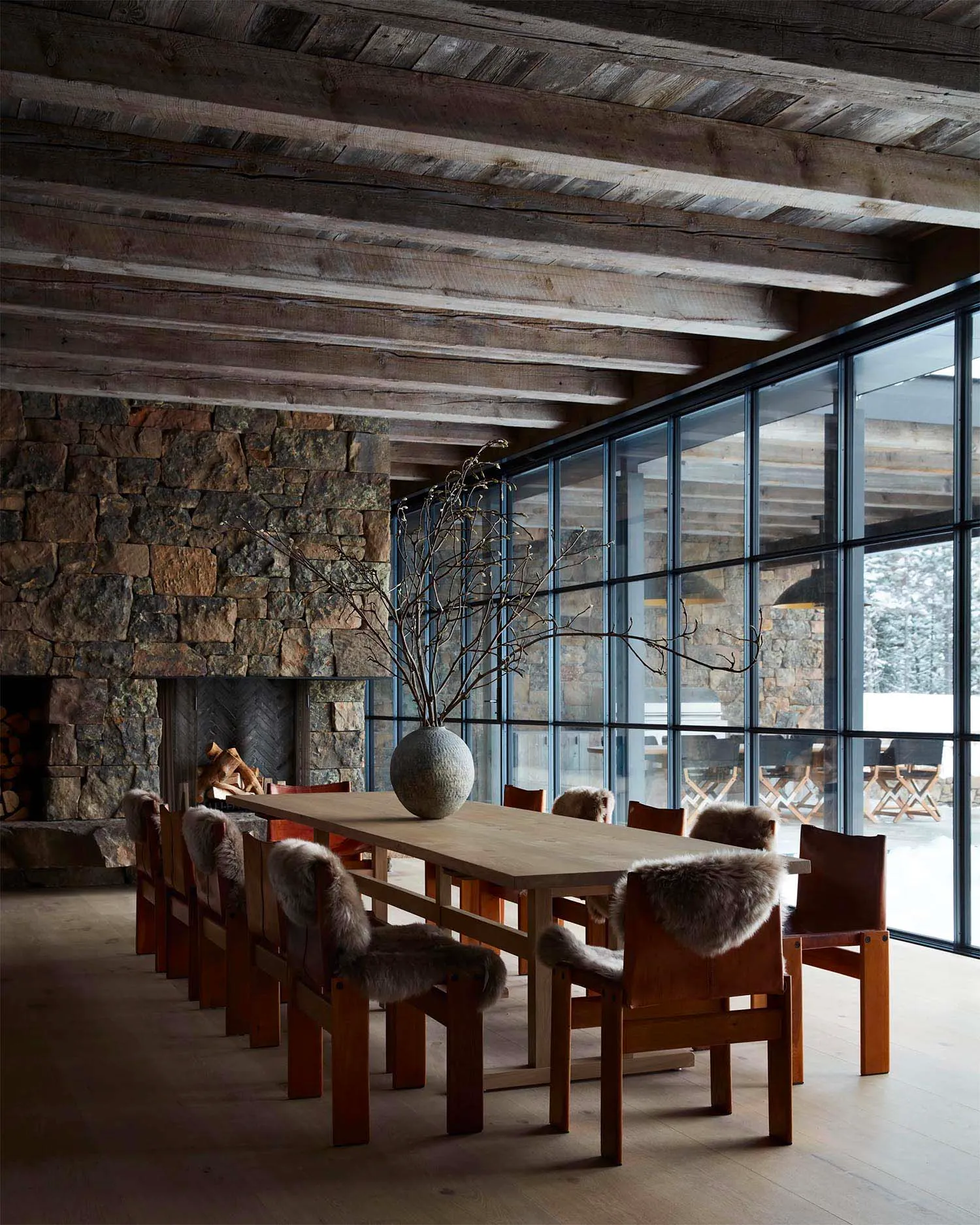 Rustic dining room featuring wooden table, fur-covered chairs, and stone accents, with large windows overlooking snowy landsc