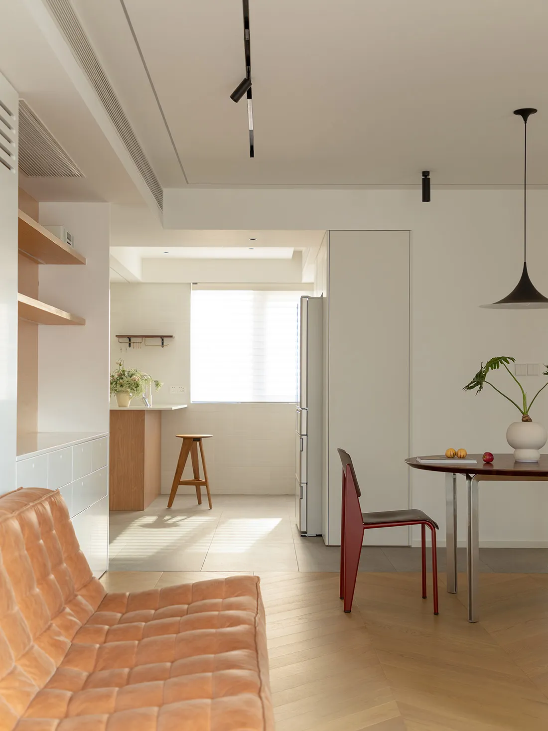 A minimalist white space featuring a wooden bench, a red chair, a round wooden table, and a wooden counter with a stool and a