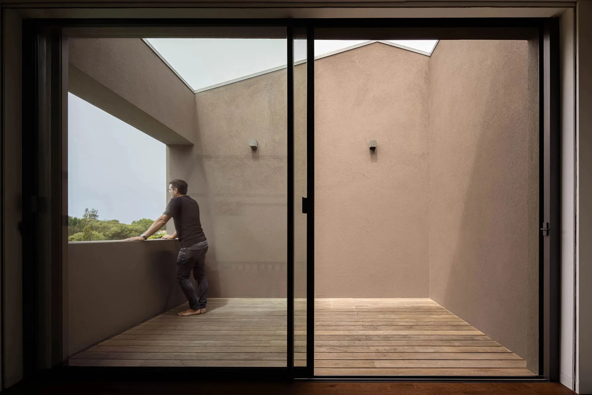 A minimalist patio with wooden flooring and concrete walls, featuring a person standing at the railing looking out.