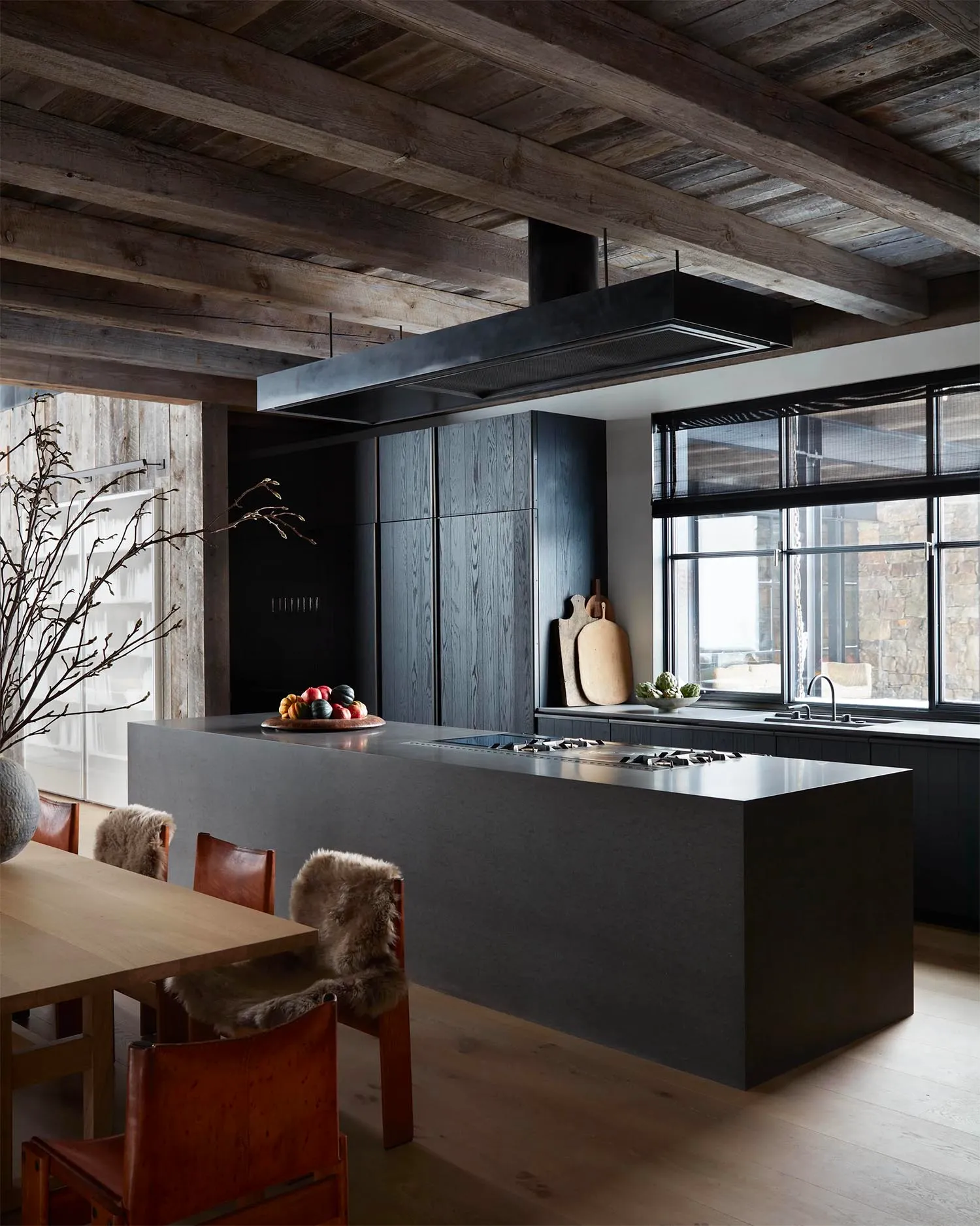 A modern minimalist kitchen featuring black cabinets, wood beams, and a dining table with a fur throw.