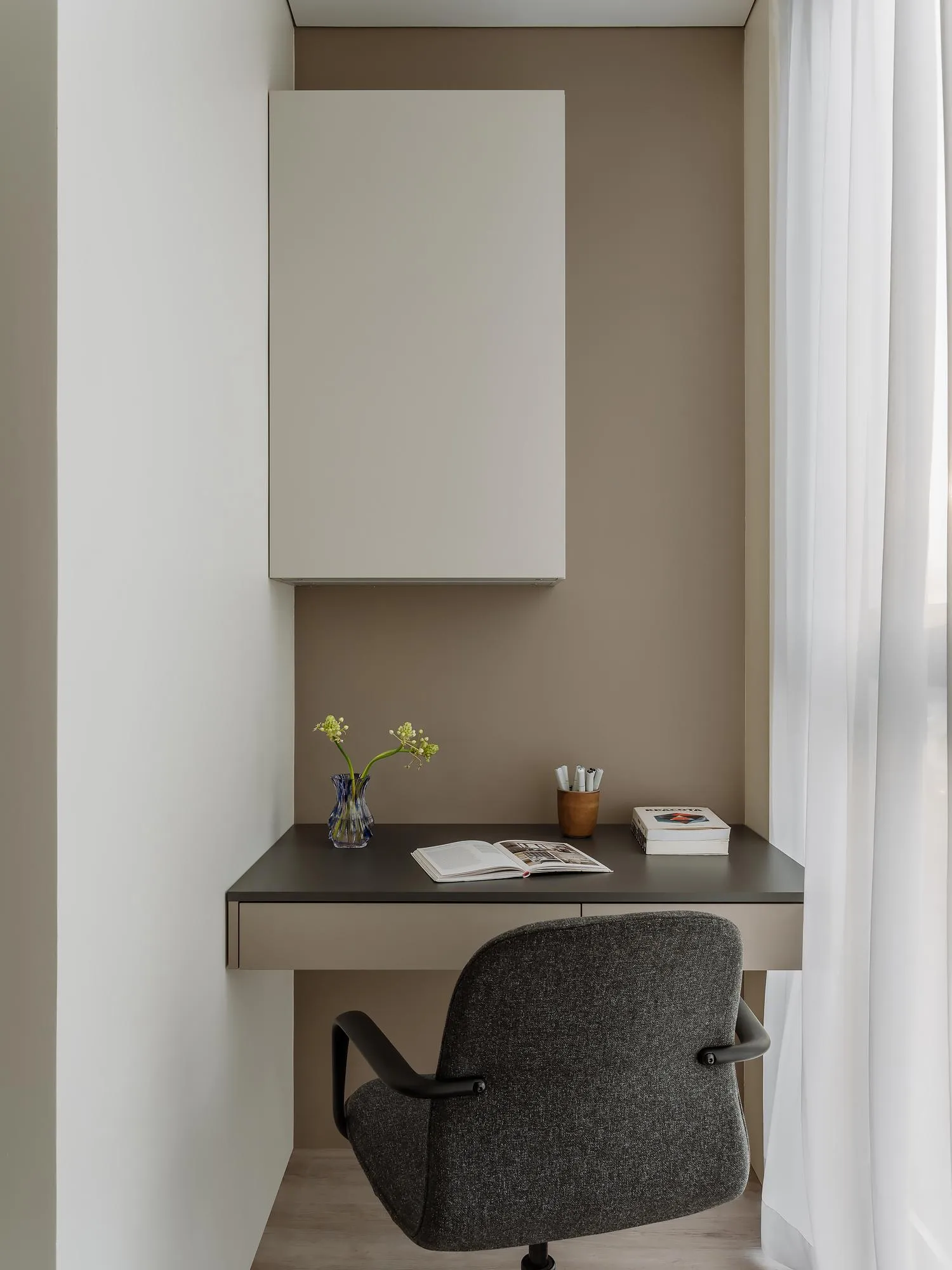 A minimalist home office featuring a beige-grey desk with a wooden drawer, a grey upholstered chair, and a beige-grey cabinet