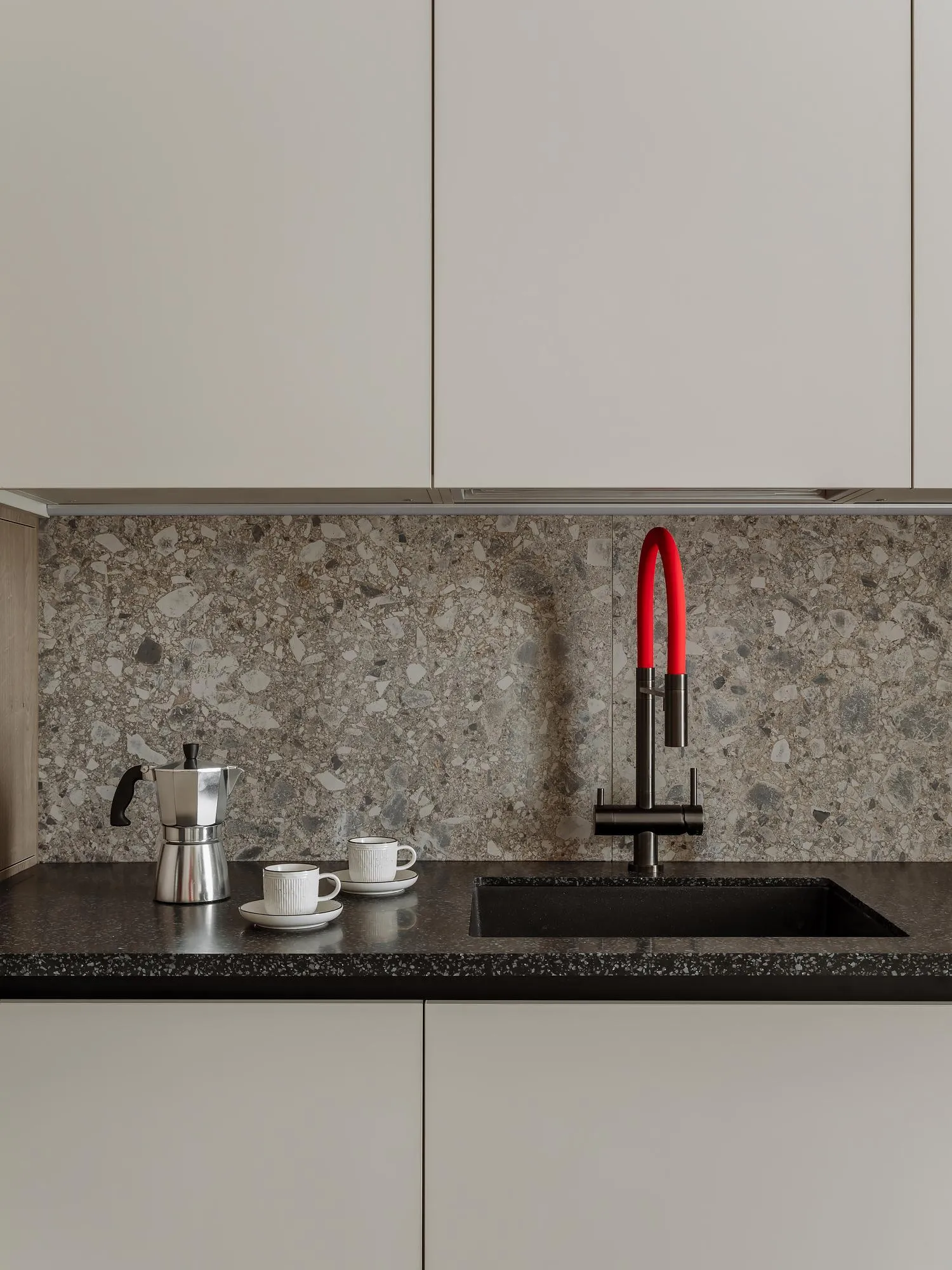A sleek modern kitchen featuring a black terrazzo countertop with a black and red faucet, white espresso cups, and a silver e