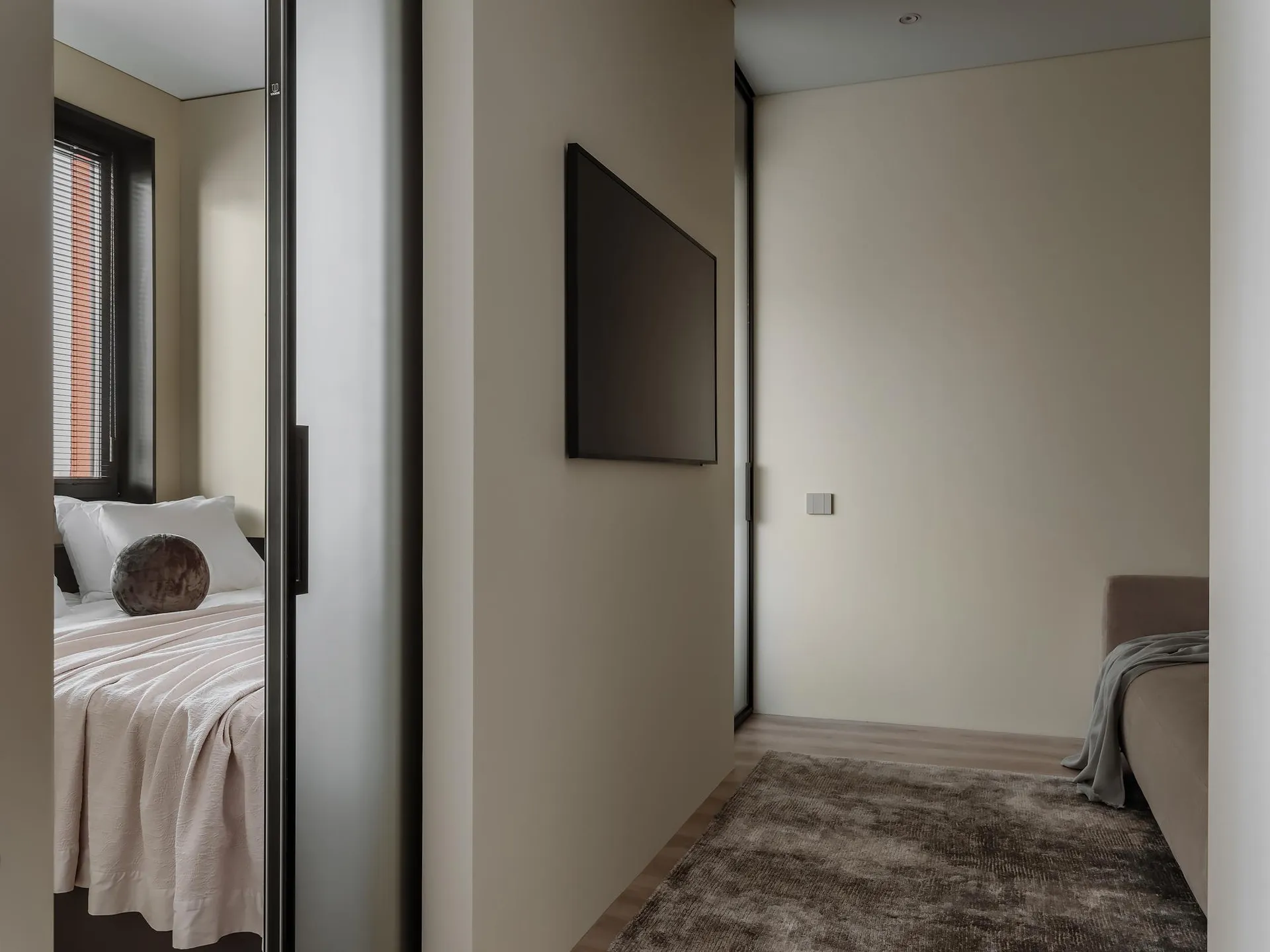 A modern bedroom with neutral-colored walls, a large flat-screen TV mounted on the wall, and a minimalistic design.