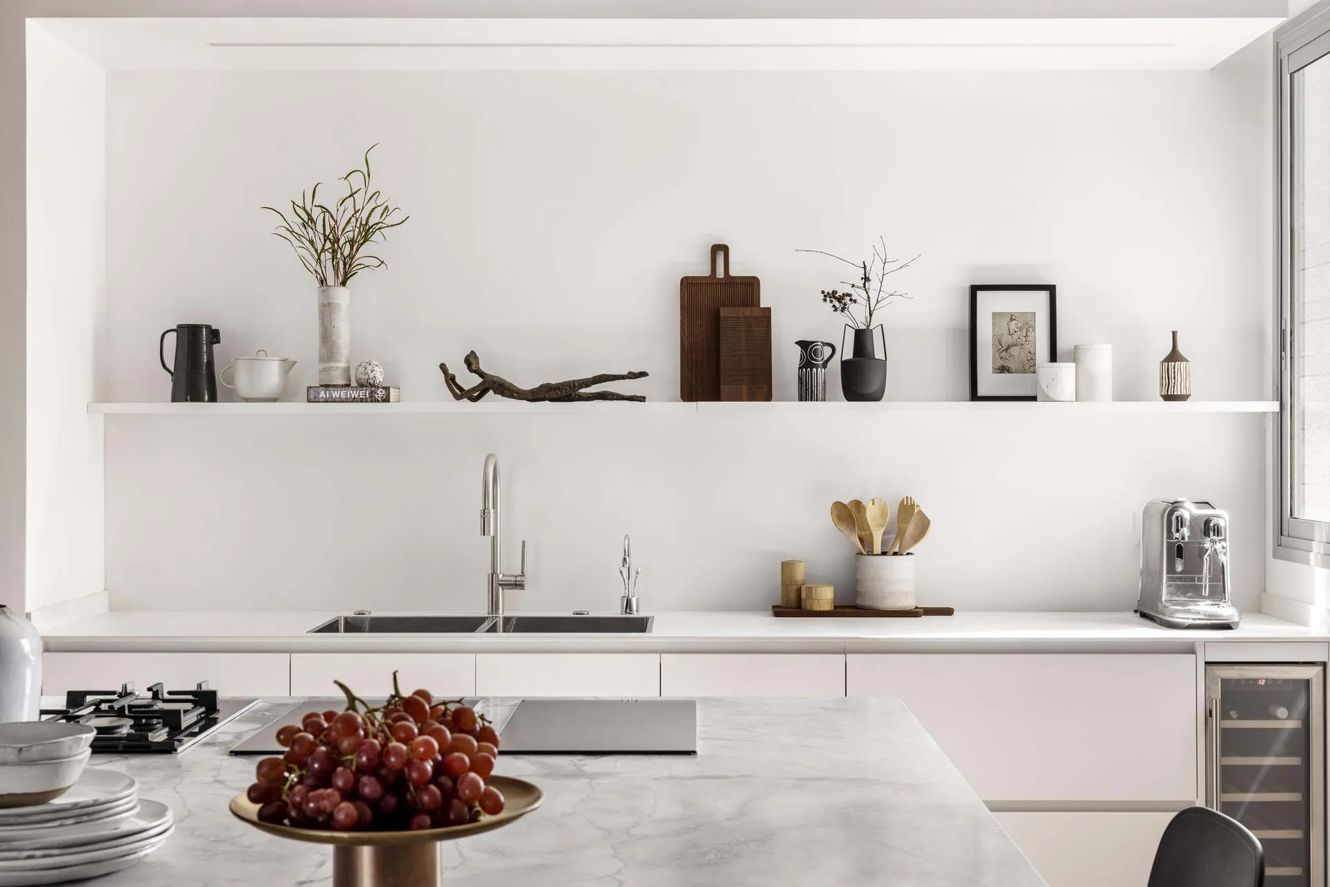 Modern minimalist kitchen featuring white marble countertops and white cabinets, with decorative items on shelves.